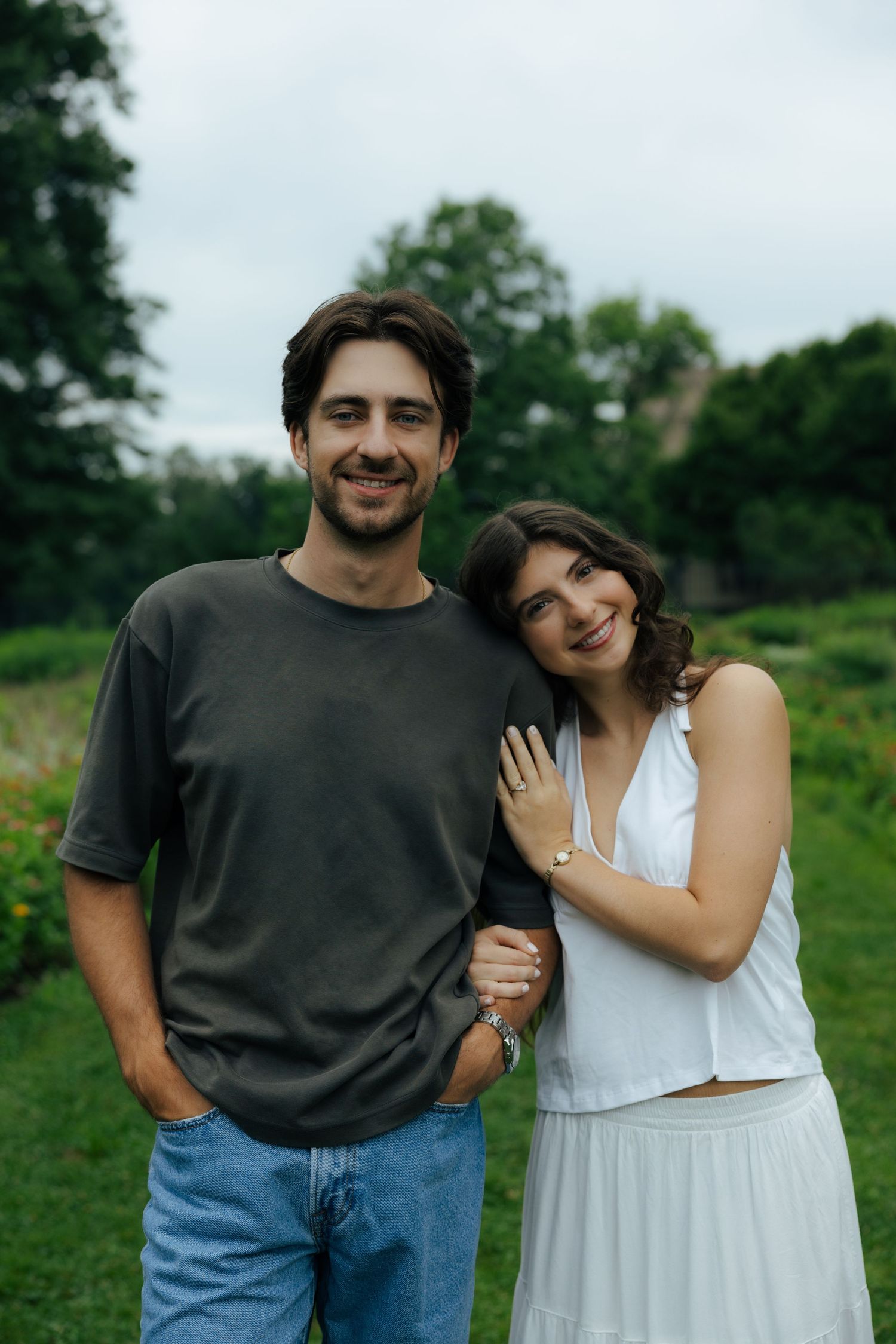 Smiling engaged couple poses together in a green park, woman in white outfit leaning on mans arm showing ring.