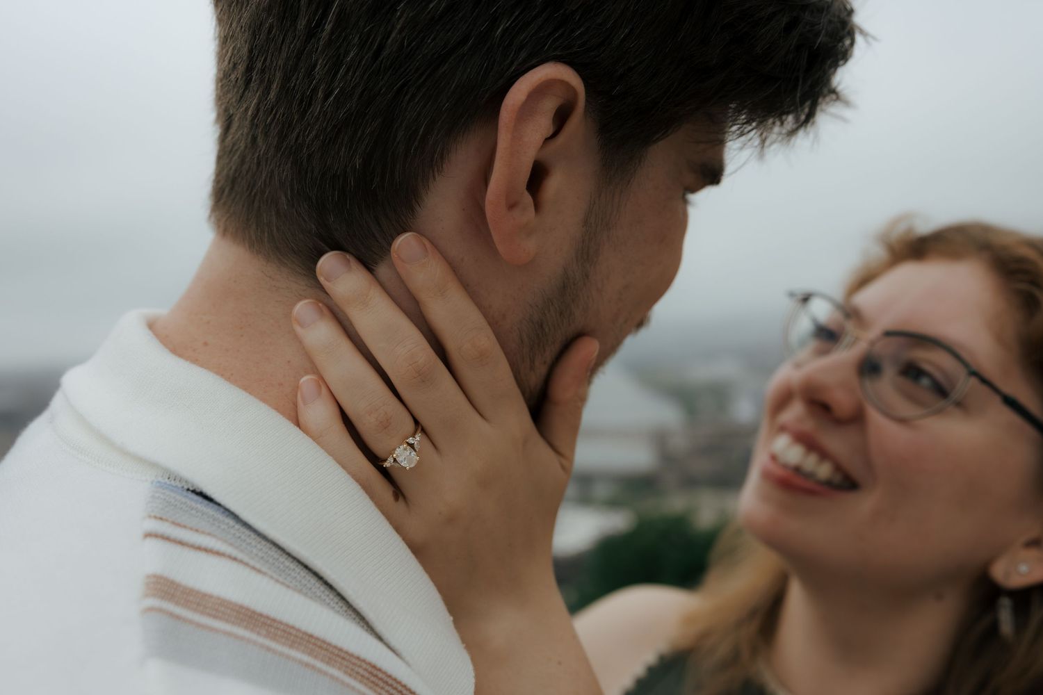 A smiling woman with glasses and an engagement ring tenderly holds a man's face during a romantic outdoor moment.