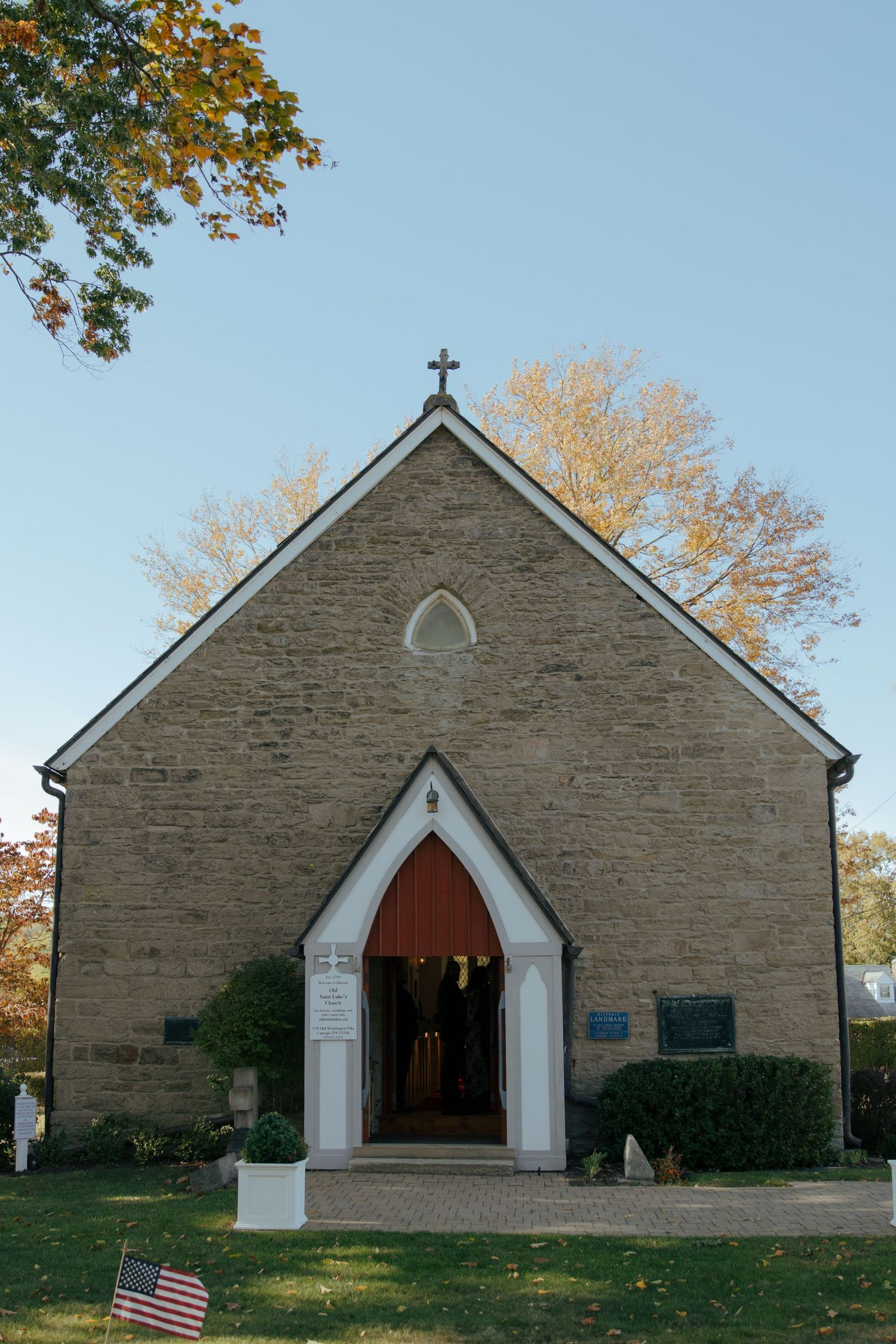 Historic stone church with Gothic arched entrance, cross atop gabled roof, red doors, American flag, surrounded by autumn trees.