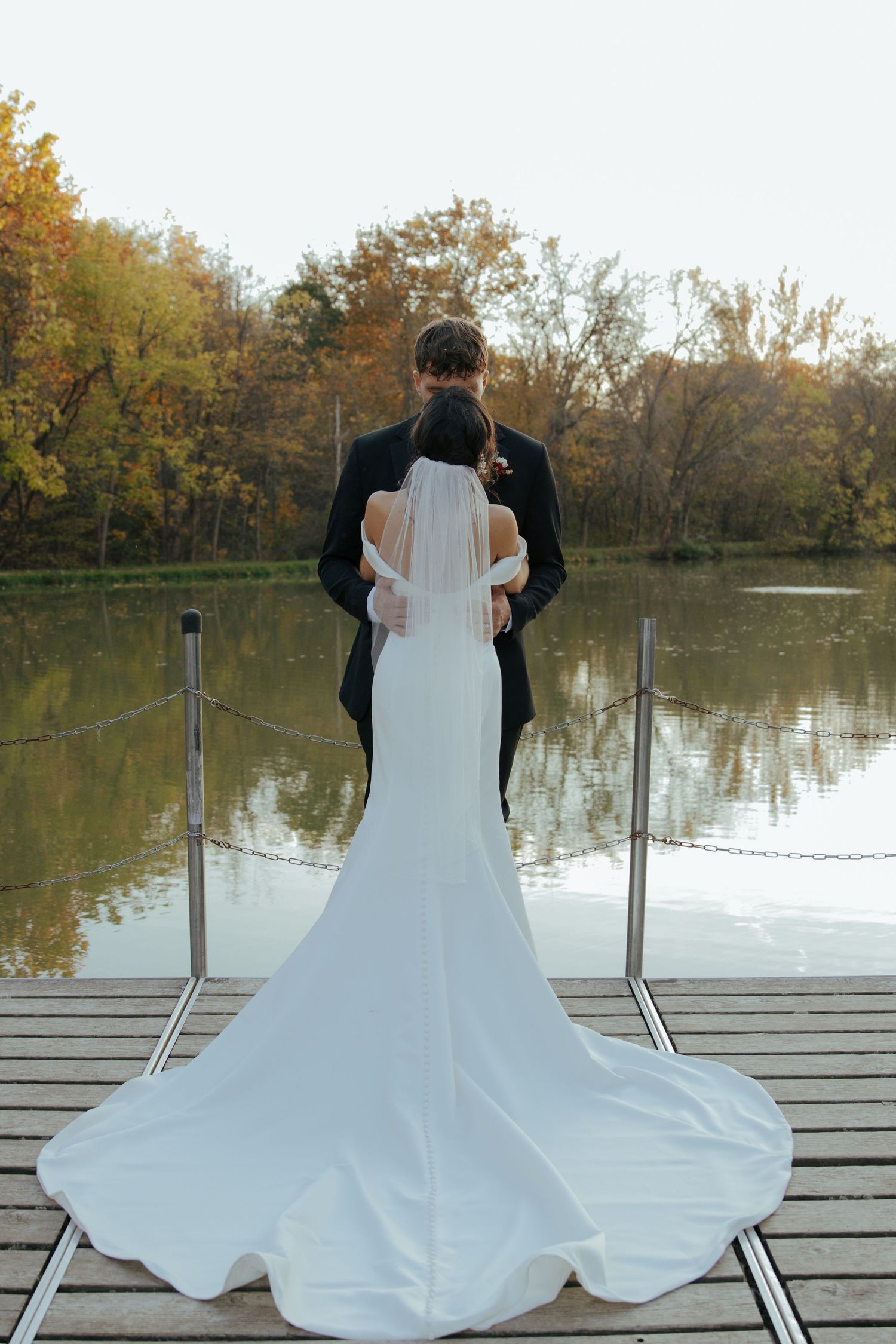 Bride and groom stand embracing on wooden dock overlooking calm lake surrounded by fall foliage.