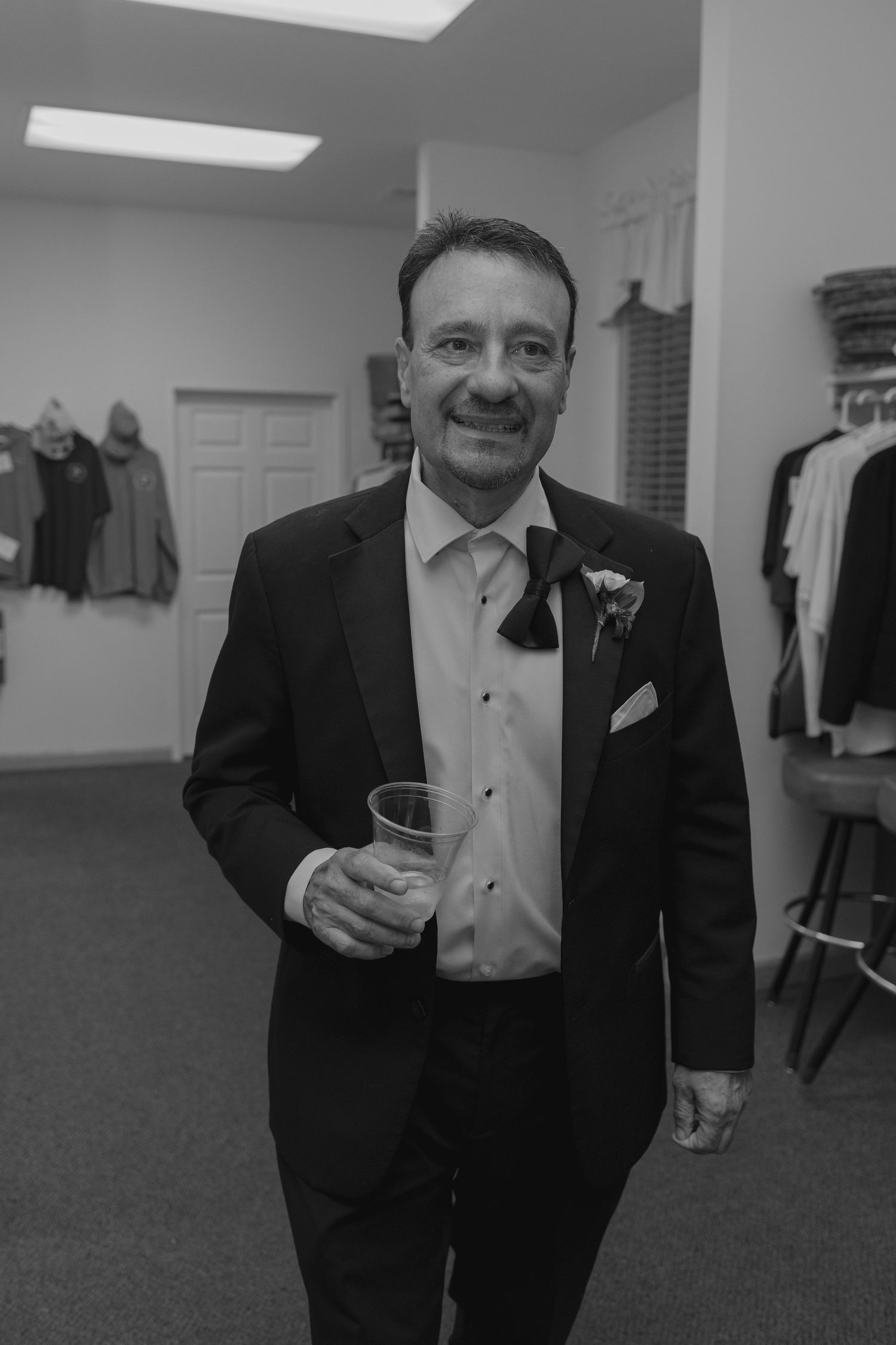 Smiling older man in a tuxedo holding a drink at a wedding reception in a black and white photo.