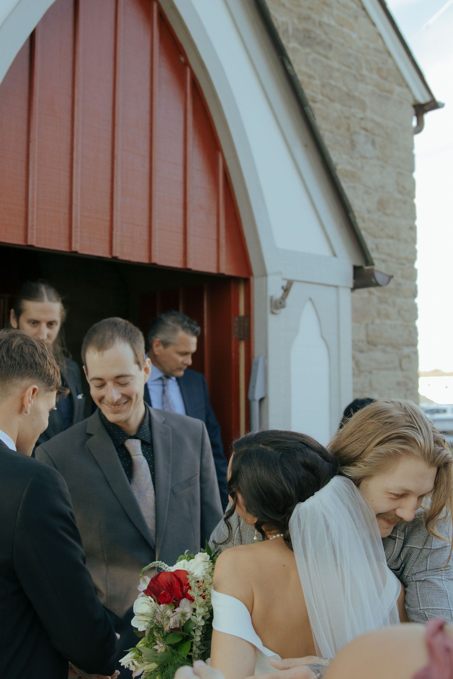 Bride and groom greet smiling wedding guests outside a charming stone church with red arched doors.