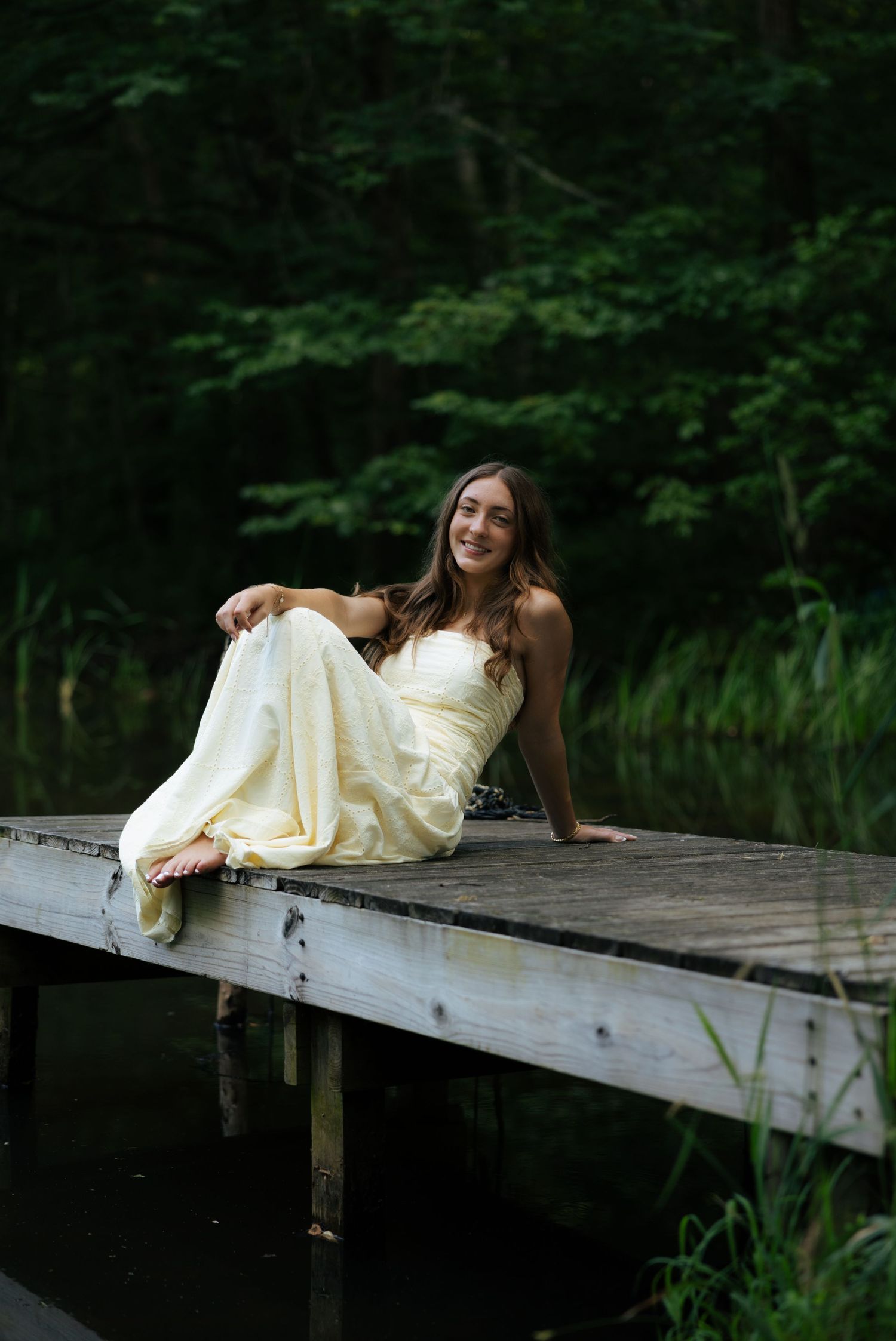 Smiling young woman in a yellow strapless dress sitting on a wooden dock beside a calm forest pond.