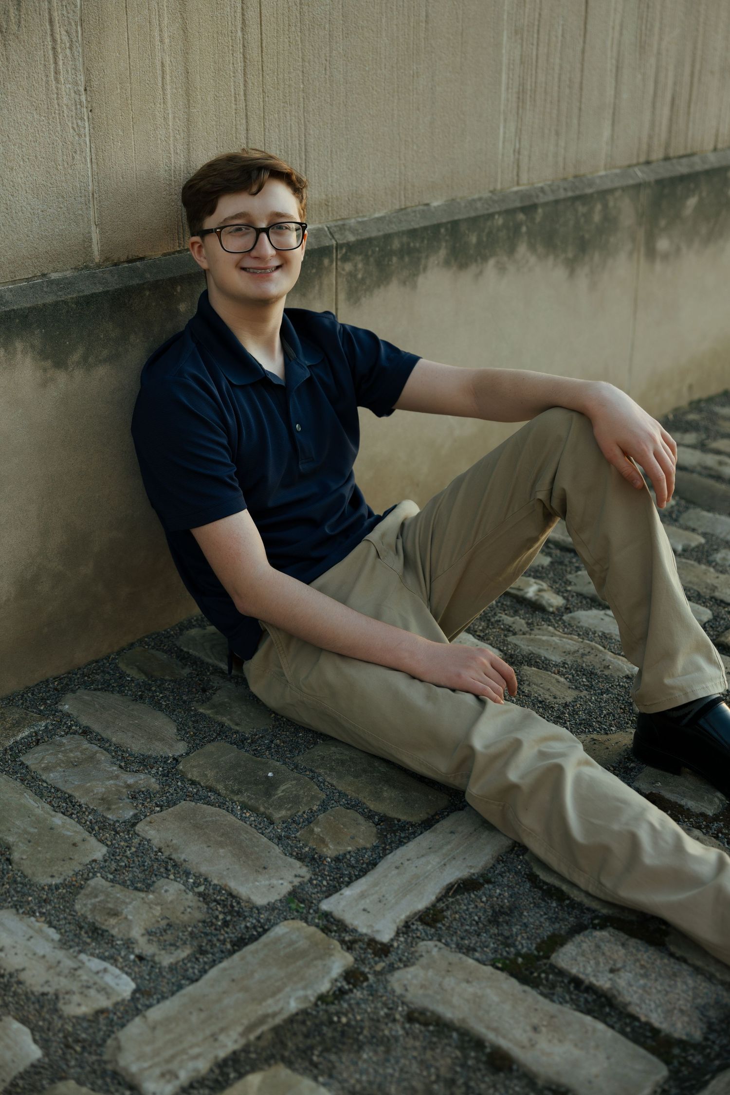 Teenage boy in navy polo and khakis sitting relaxed against concrete wall on cobblestone surface.