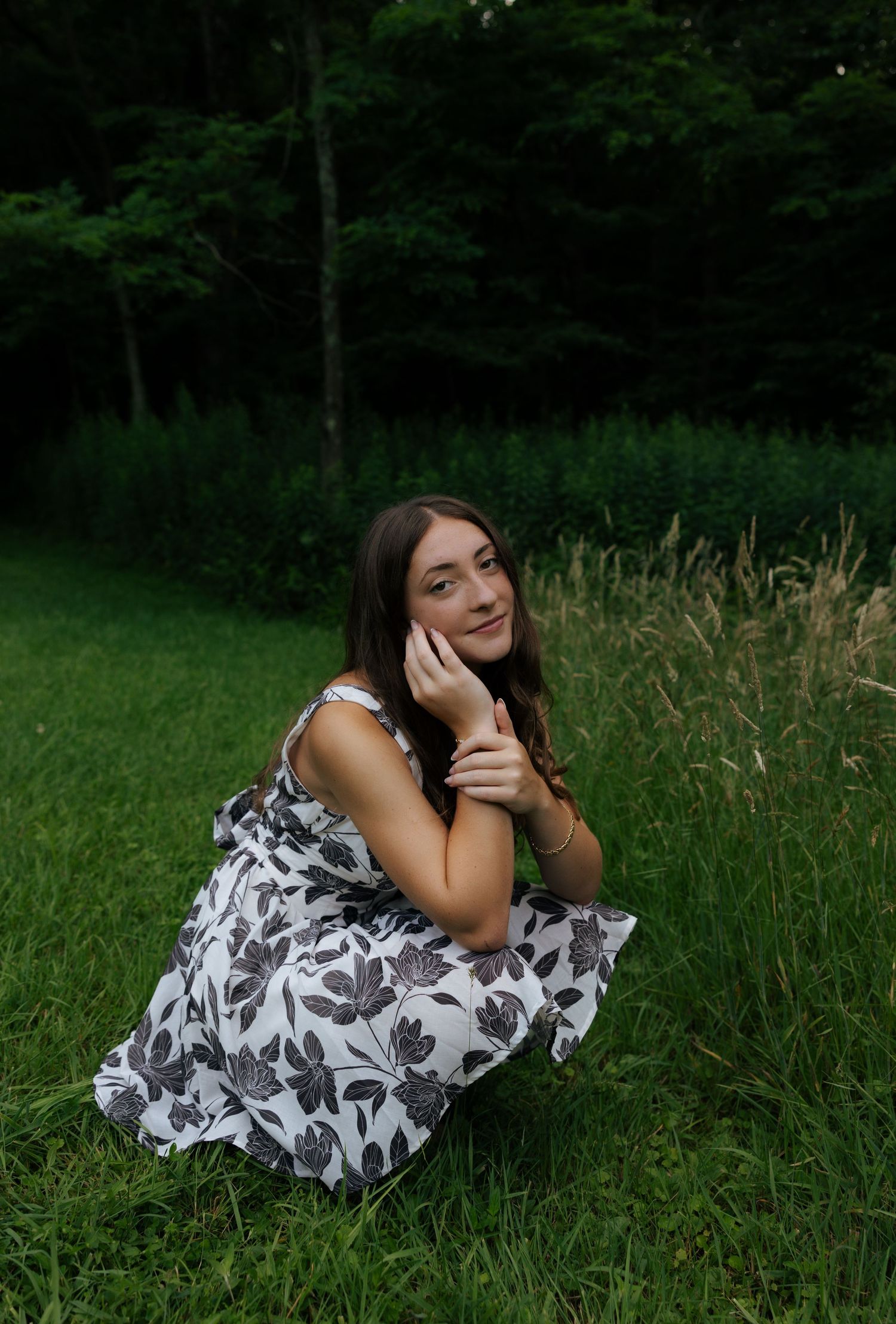 Young woman in white floral dress sitting on green grass, smiling at camera in park.