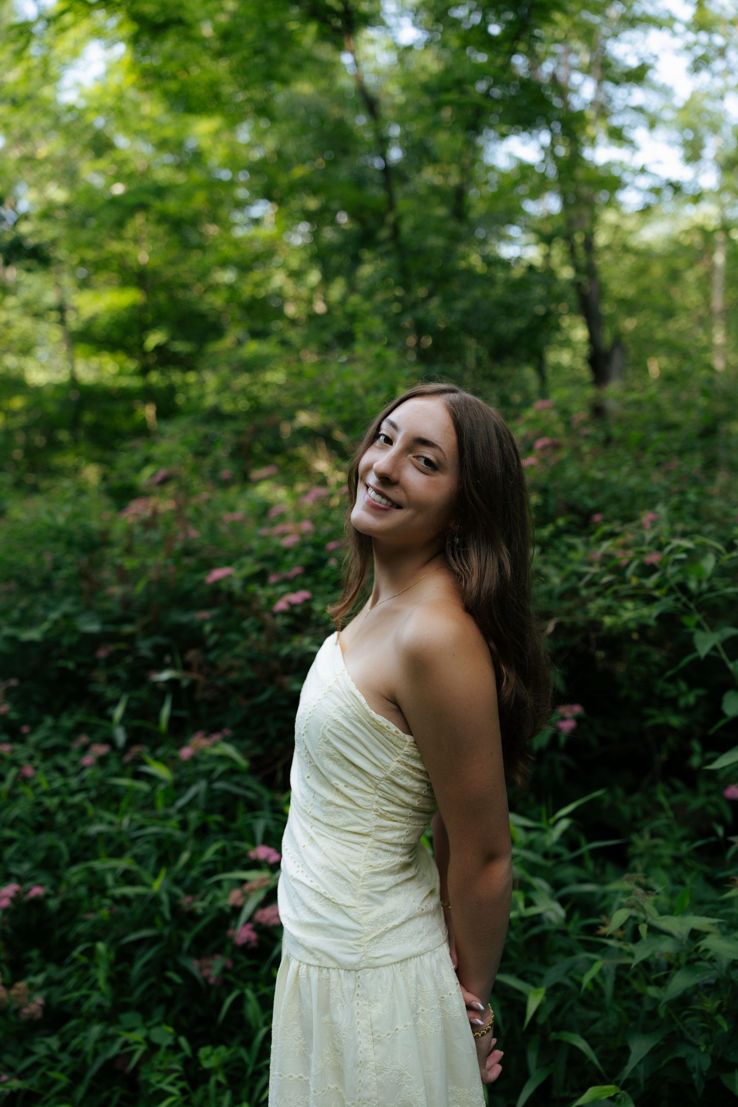 Smiling young woman in white strapless dress posing among lush green forest foliage.