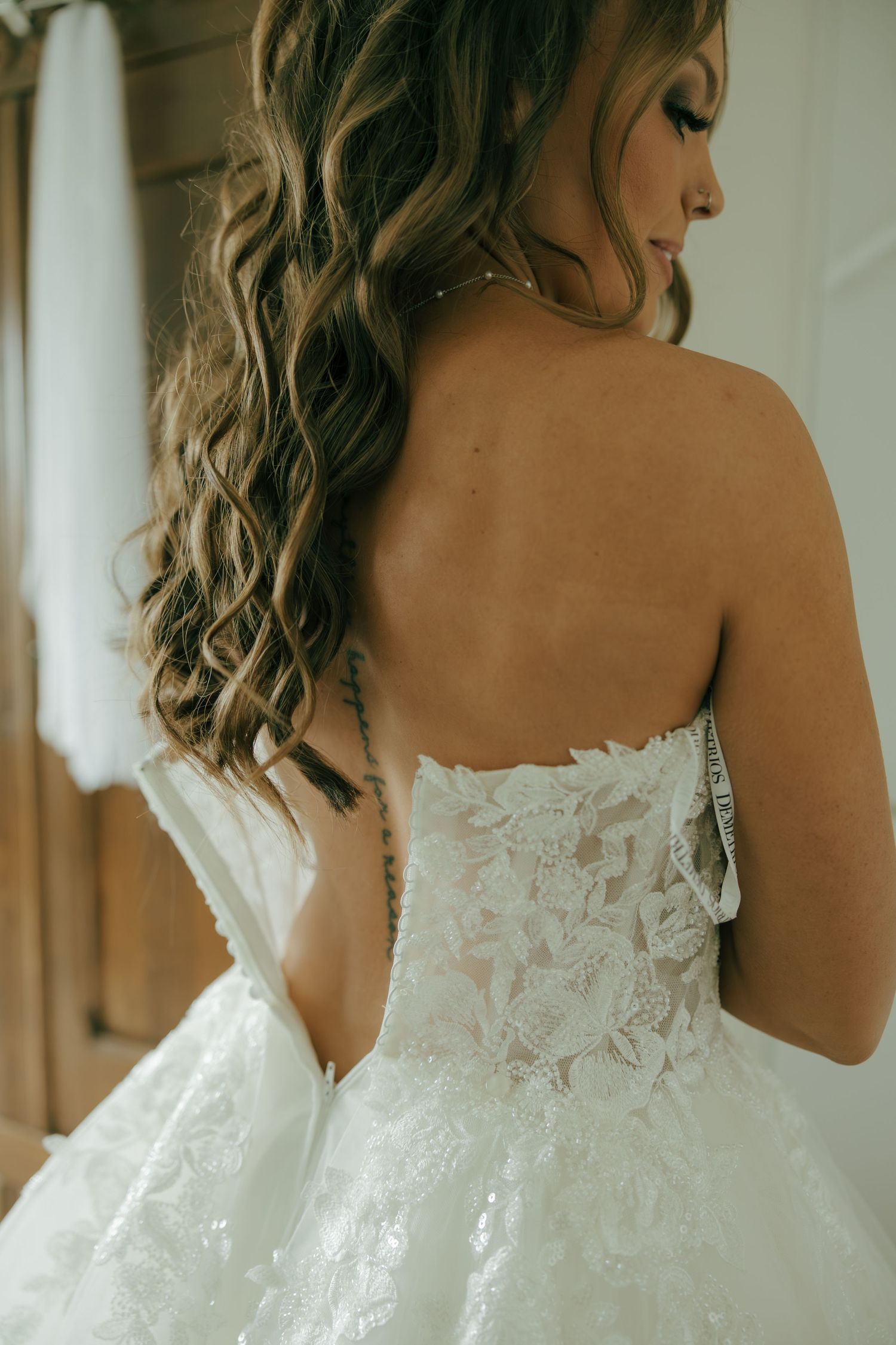 Bride in strapless lace wedding gown showing intricate back detail with flowing curly hair.