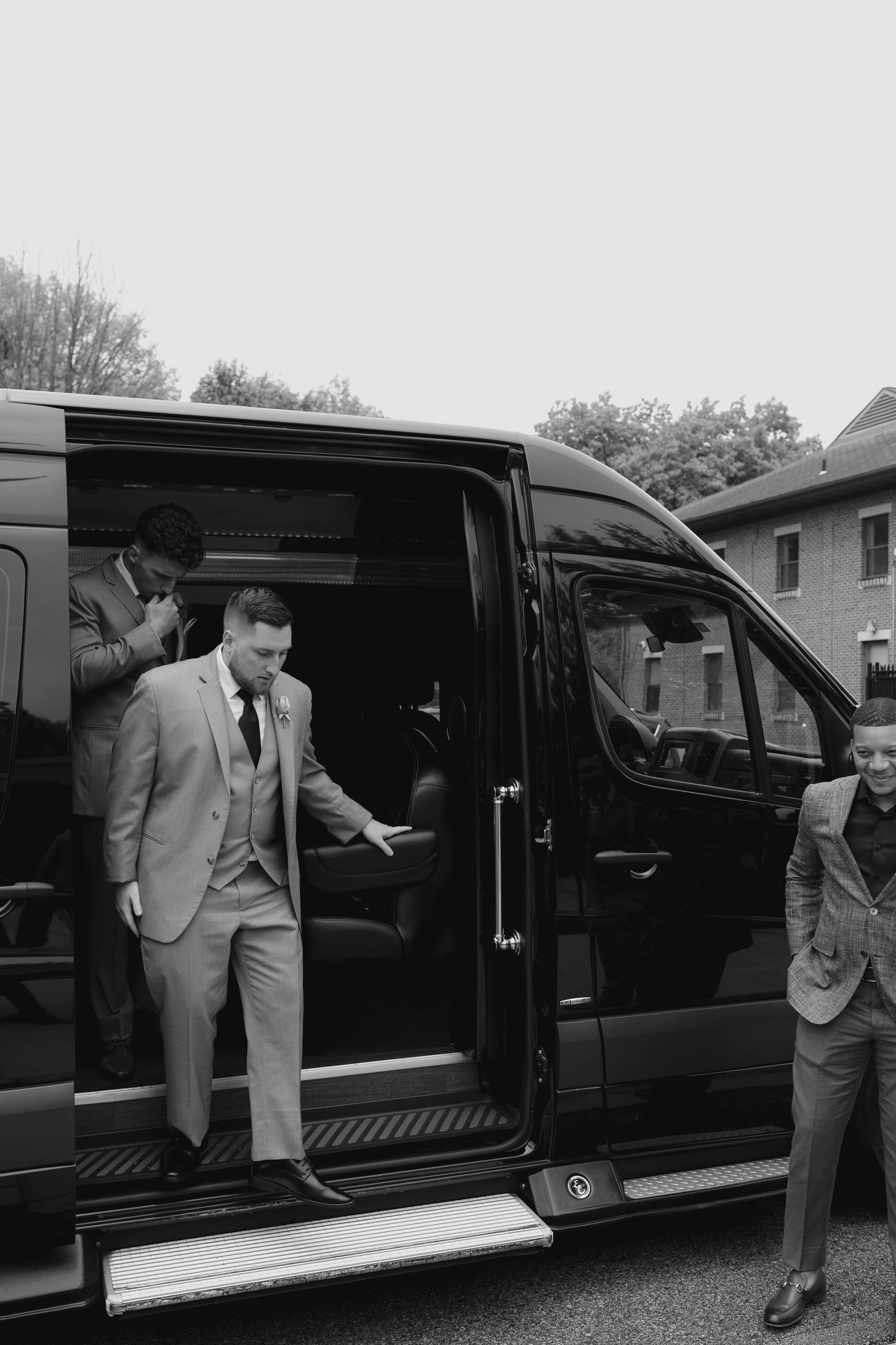 Groomsmen in gray suits stepping out of a black luxury van at a wedding venue.