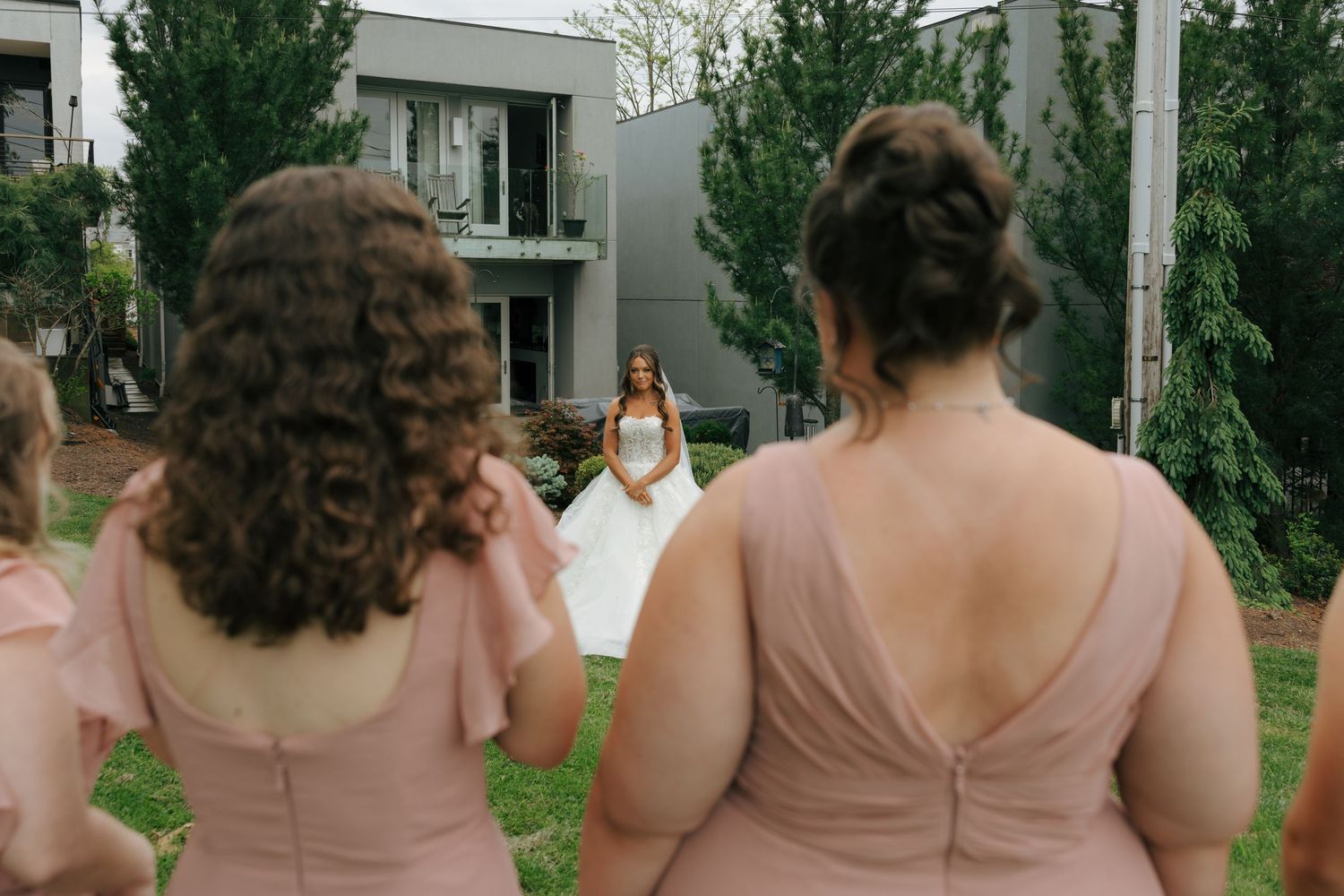 Bridesmaids in blush pink dresses watch a bride in a white gown walk toward them outdoors.
