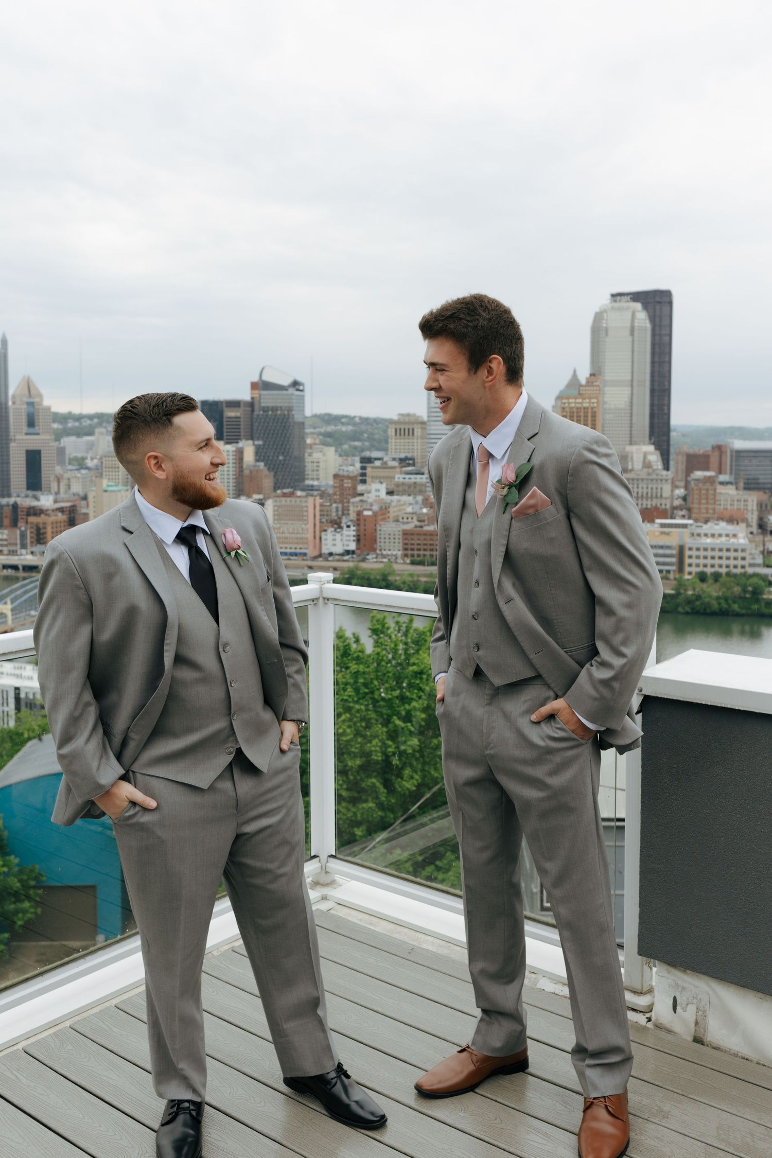 Two groomsmen in grey suits chat on rooftop with Pittsburgh city skyline behind them.