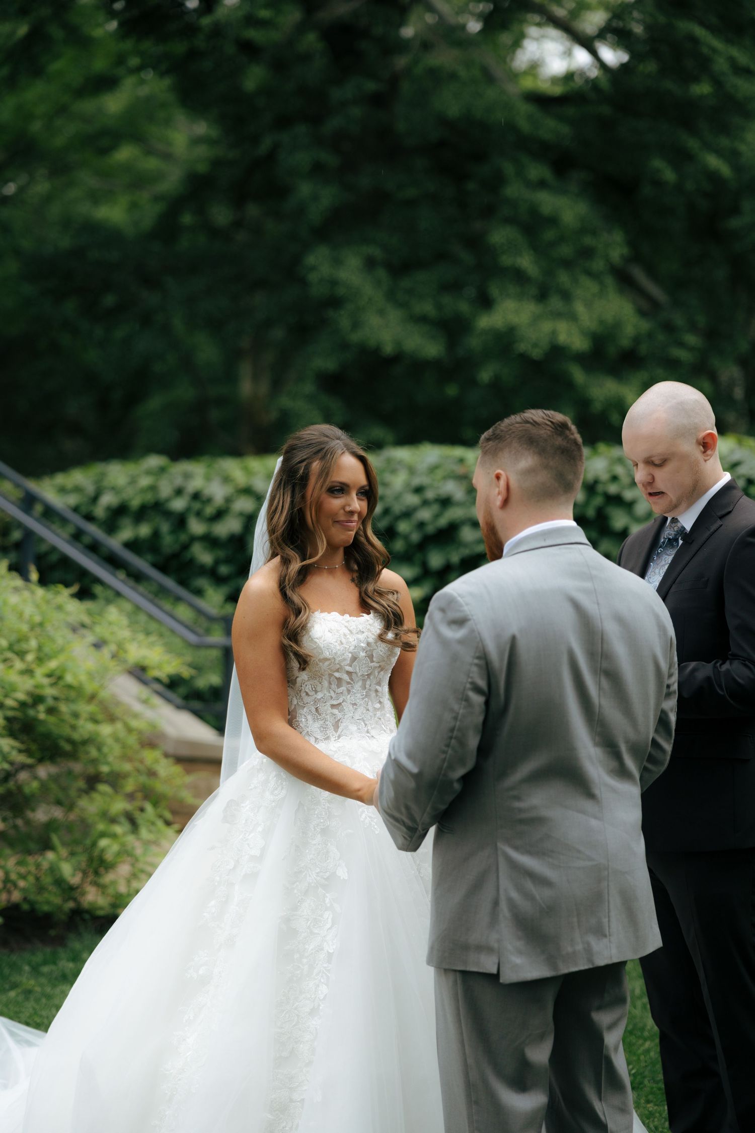 Bride in white lace ballgown smiles at groom during outdoor garden wedding ceremony with officiant.