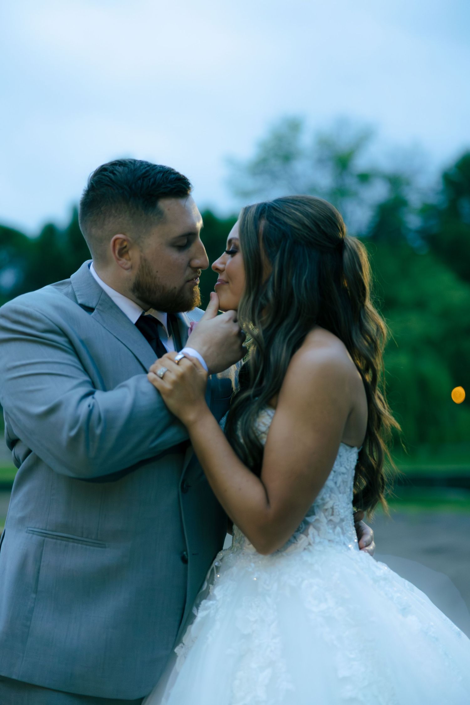 Groom in gray suit tenderly touches brides chin as they share an intimate moment at dusk outdoors.