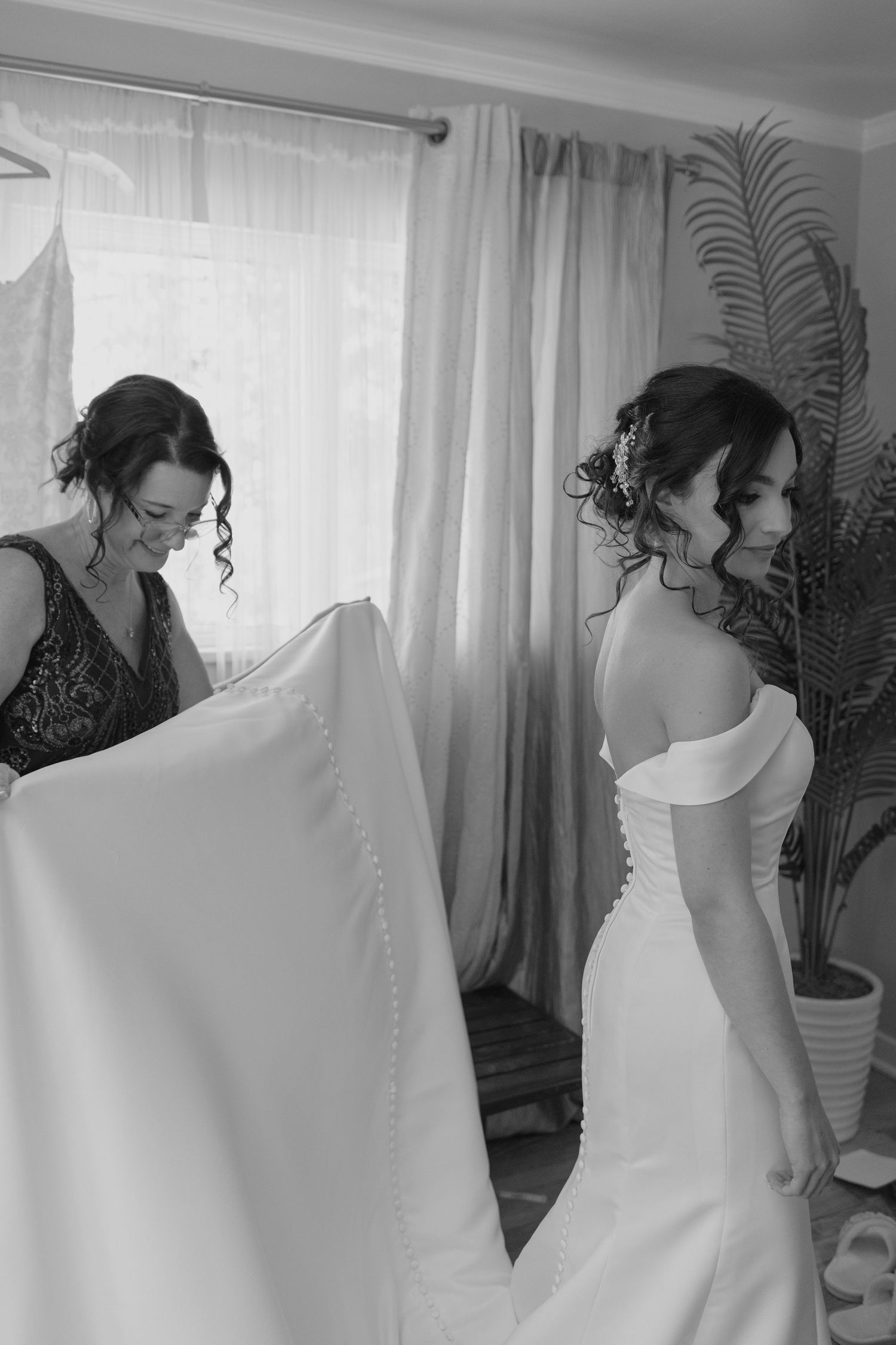 Black and white photo of mother adjusting bride's off-shoulder wedding dress train before ceremony.