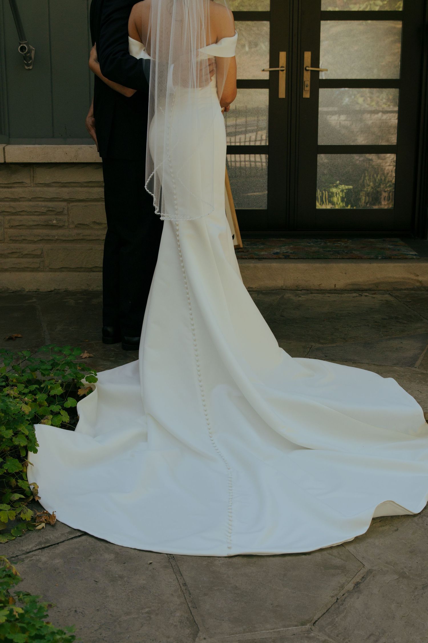 Bride in elegant off-shoulder white wedding gown with long train and veil, standing with groom at stone entrance.