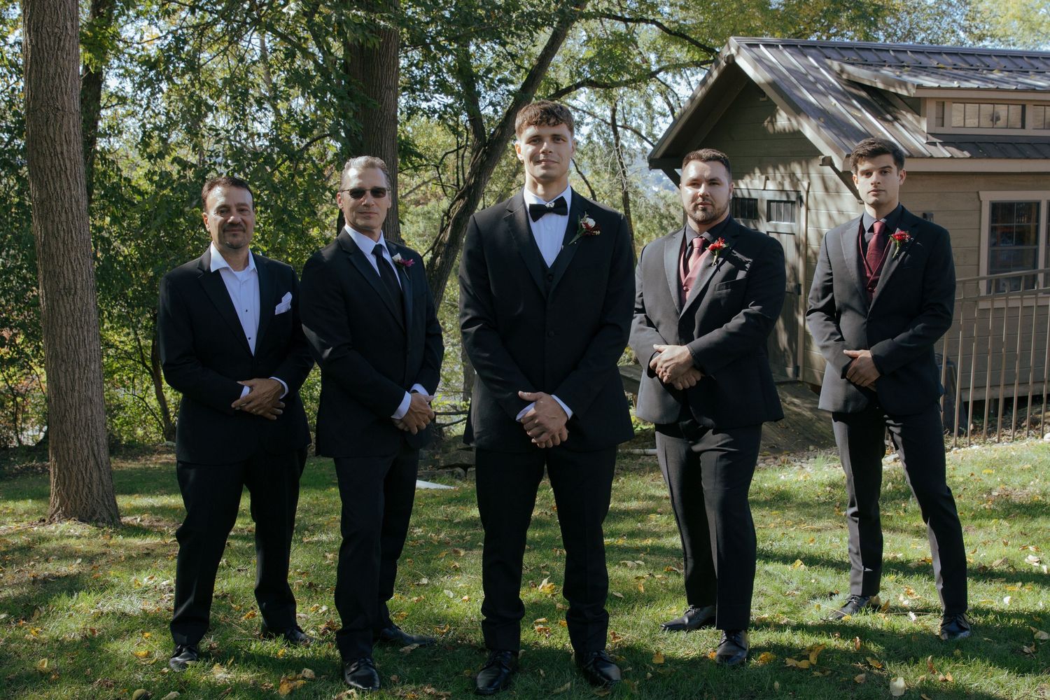 Five groomsmen in black suits pose outdoors near a rustic wooden building surrounded by green trees.