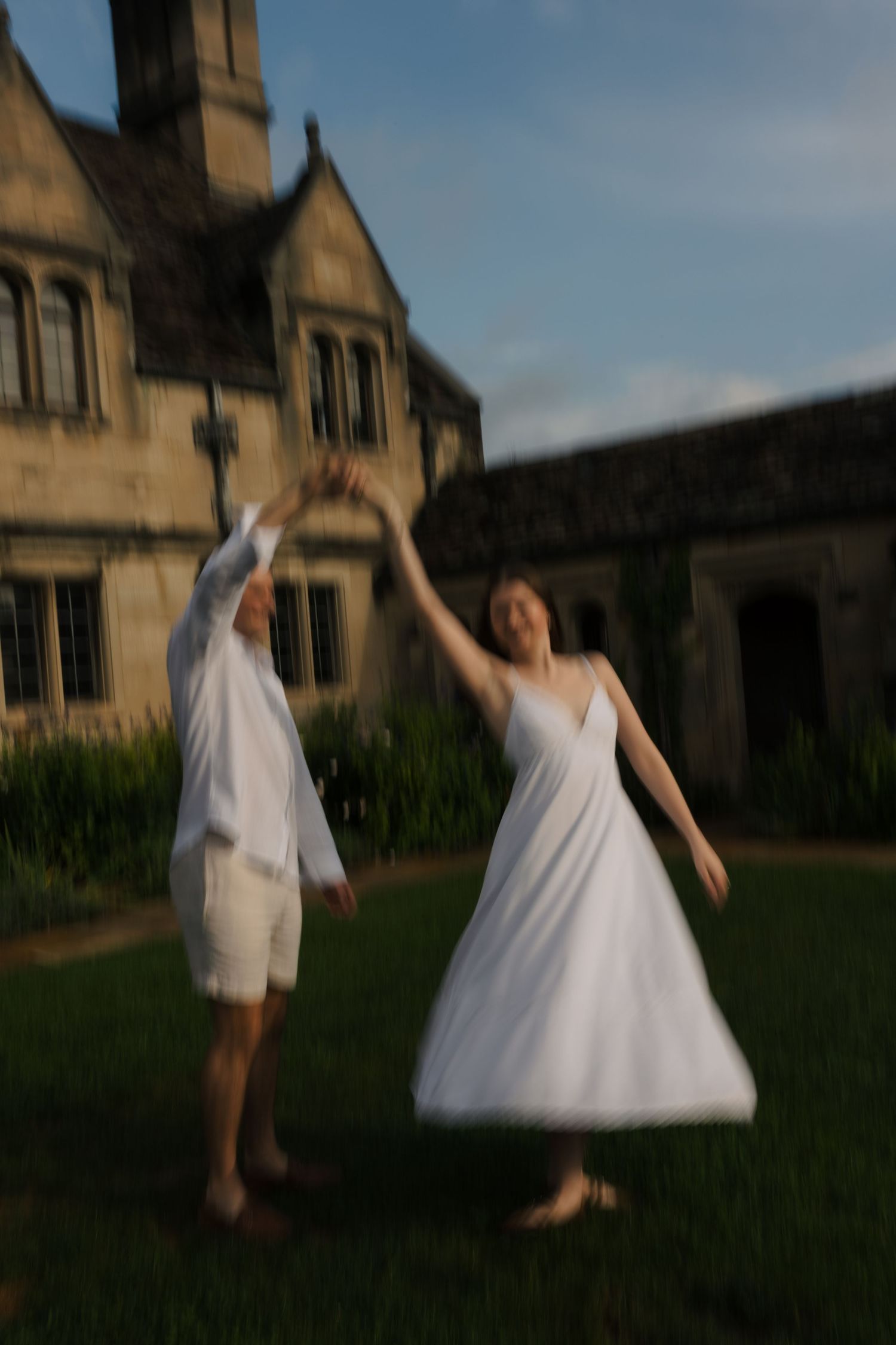 Couple in white outfits dancing joyfully on green lawn in front of a historic stone manor house.