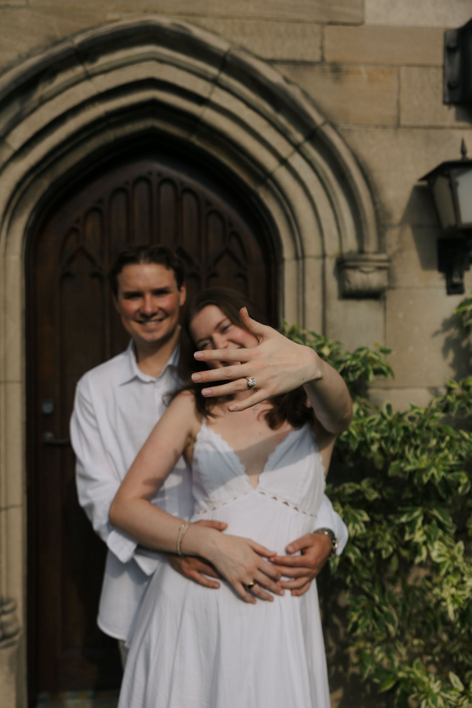 Engaged couple in white posing before Gothic arch doorway, showing ring.