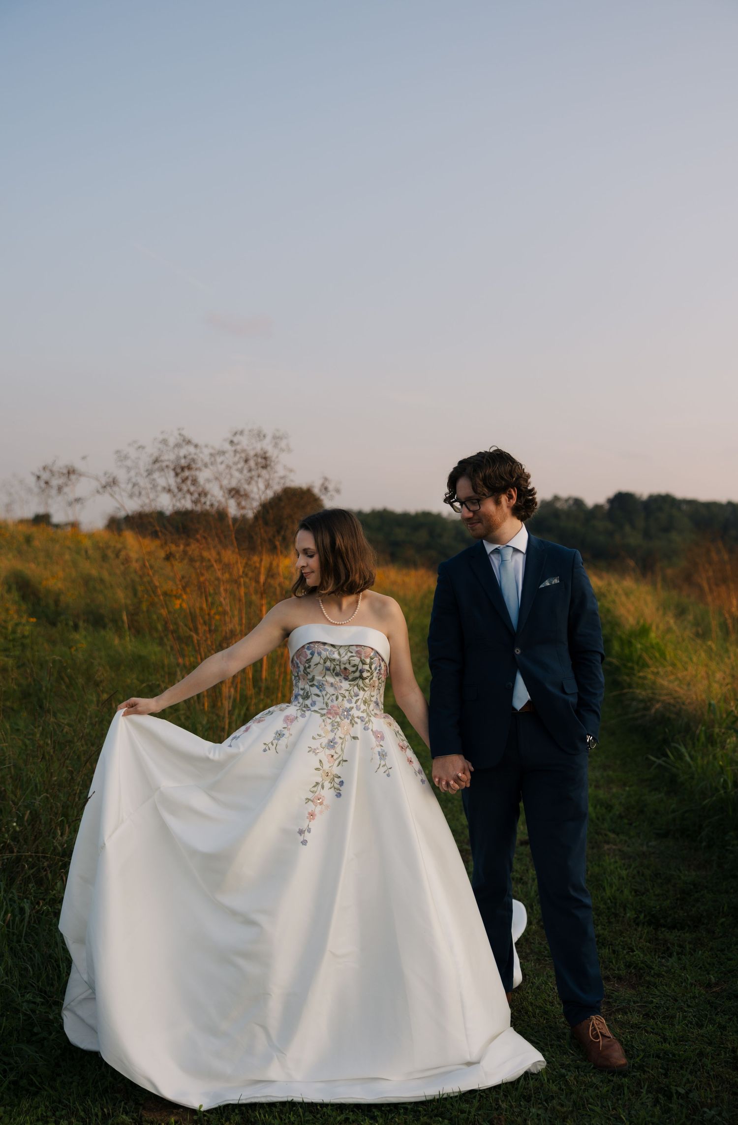 Bride in floral embroidered white gown holds dress while walking hand-in-hand with groom in navy suit through golden meadow.