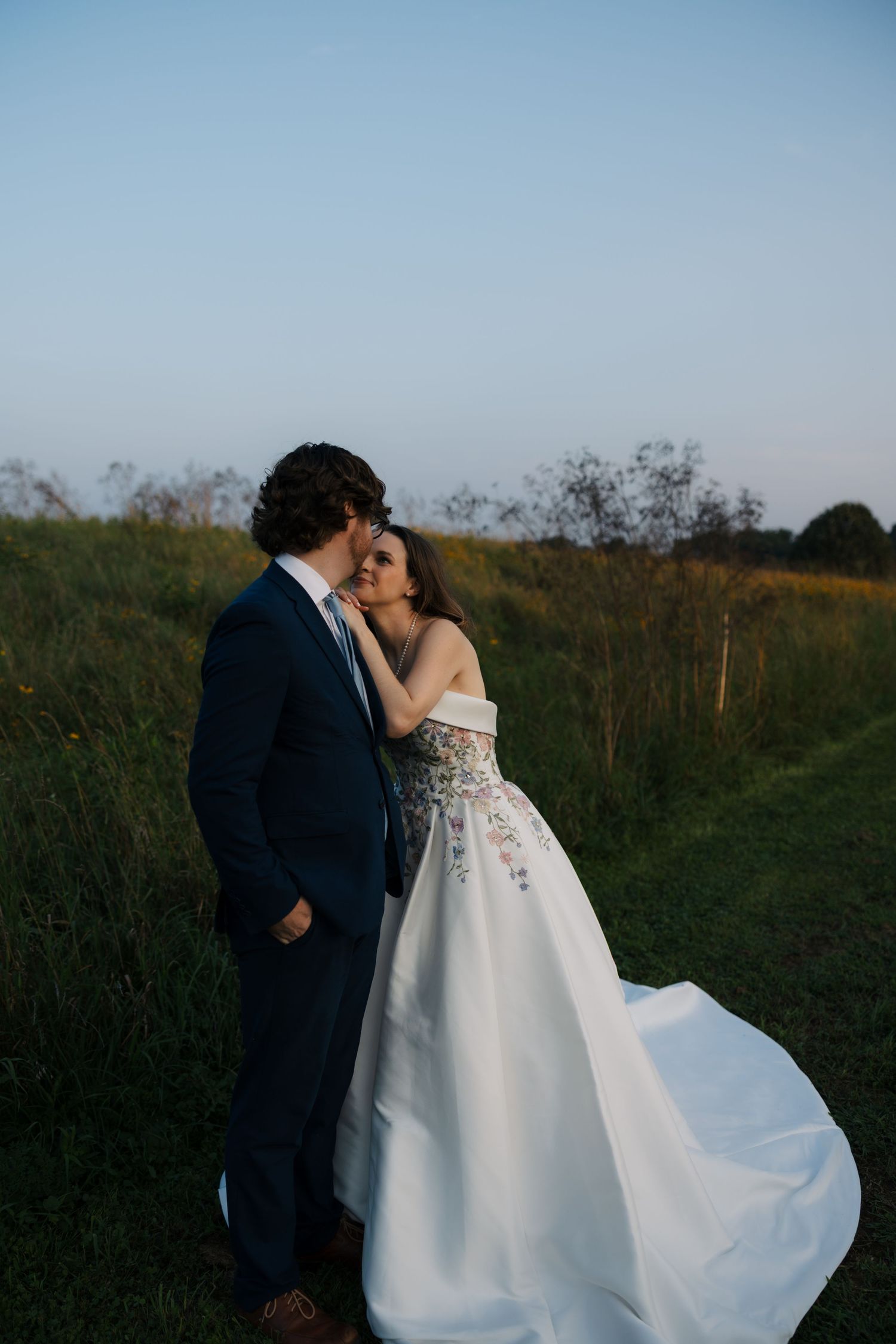 Bride in flowing floral off-shoulder ballgown embraces groom in navy suit in a lush green meadow at dusk.
