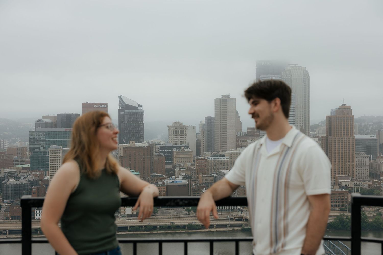 Two young adults chat on a rooftop overlooking a city skyline on a cloudy day.