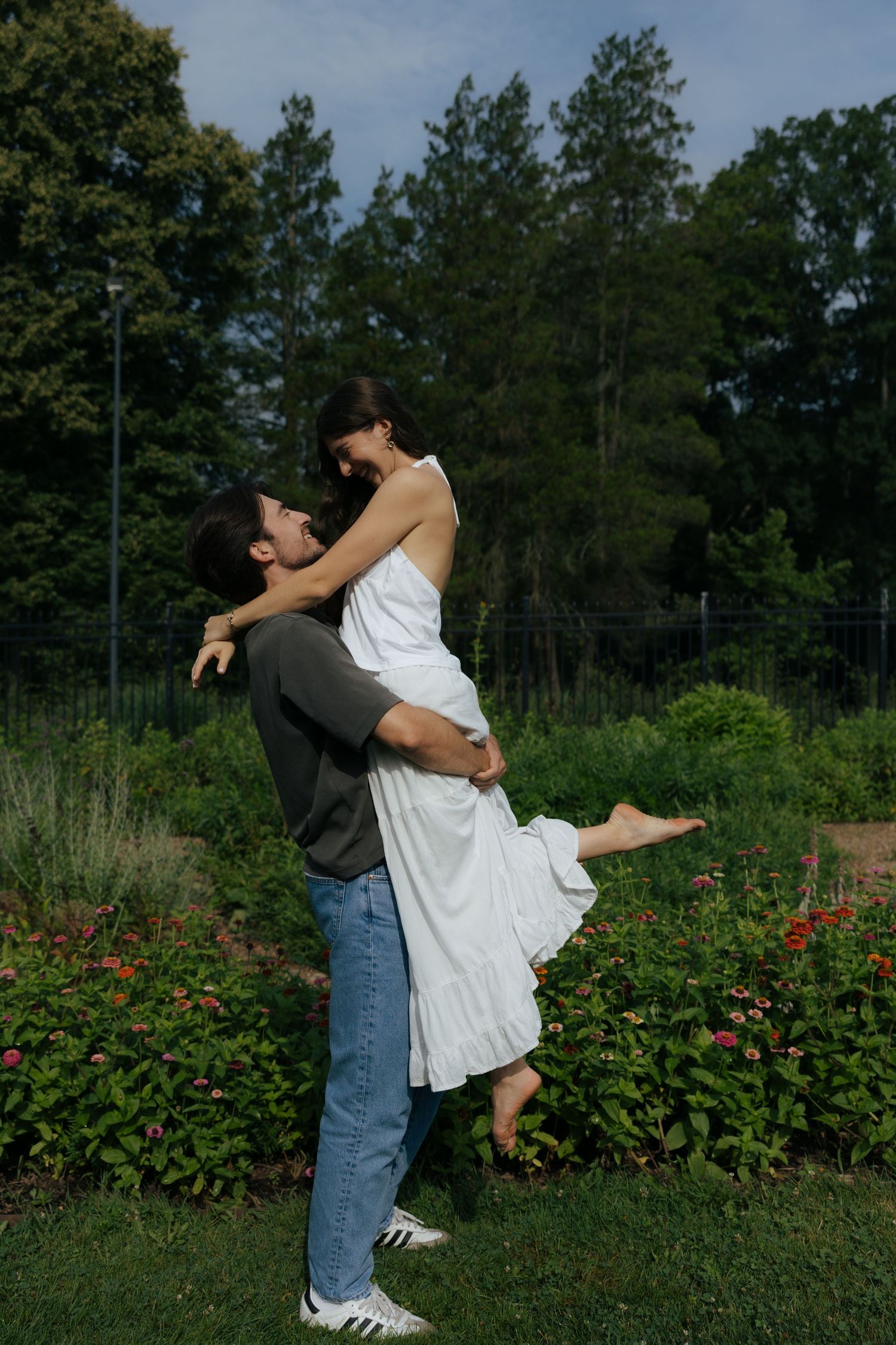A joyful man lifts a woman in a white dress high in the air in a colorful garden on a sunny day.