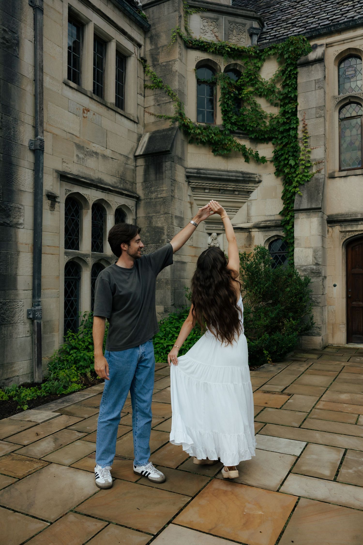 A couple dances together in a courtyard of a historic ivy-covered stone building, man spinning woman in white dress.