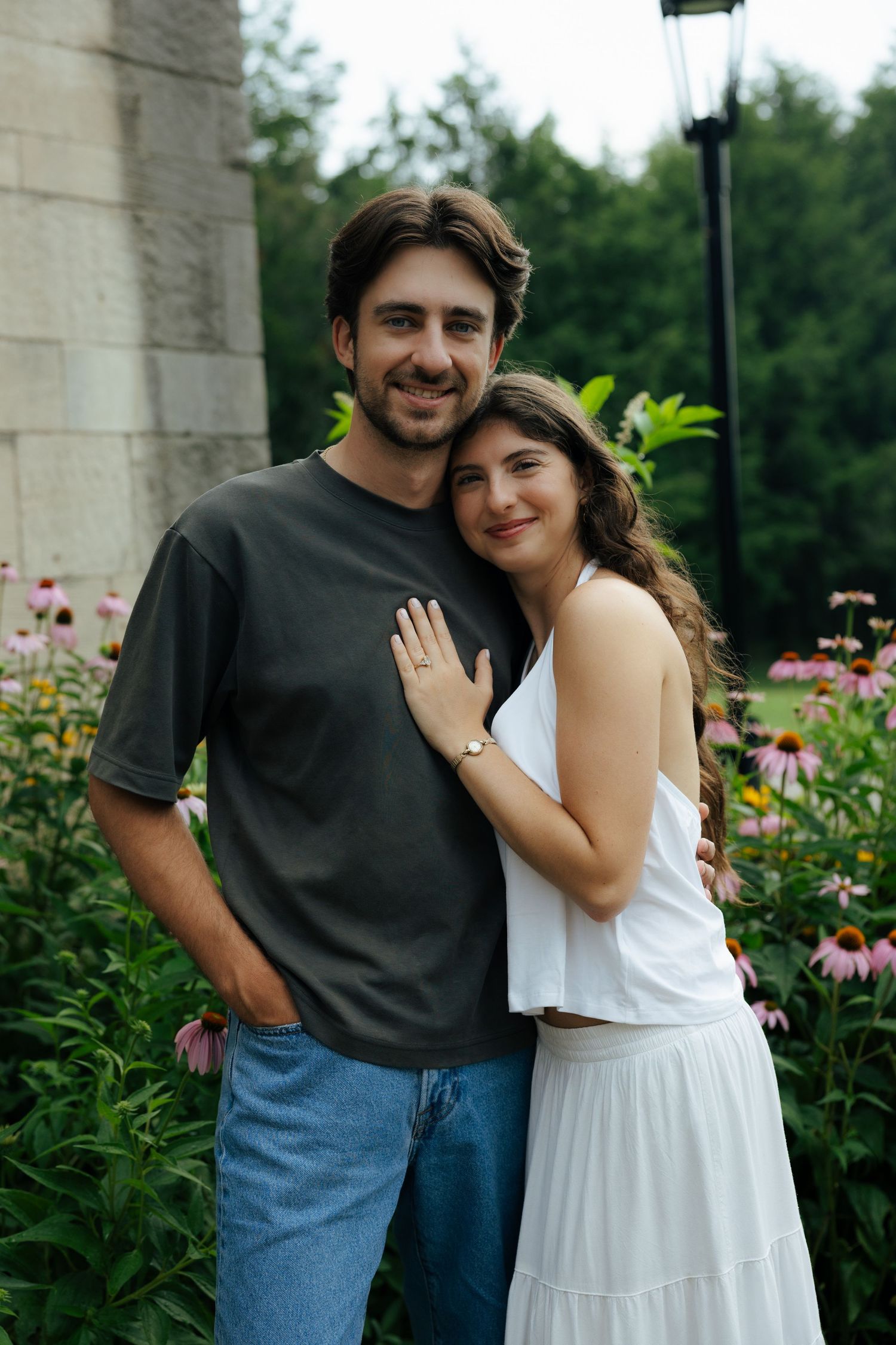 A smiling couple embraces outdoors near blooming pink echinacea flowers, the woman in a white dress leaning on the man.