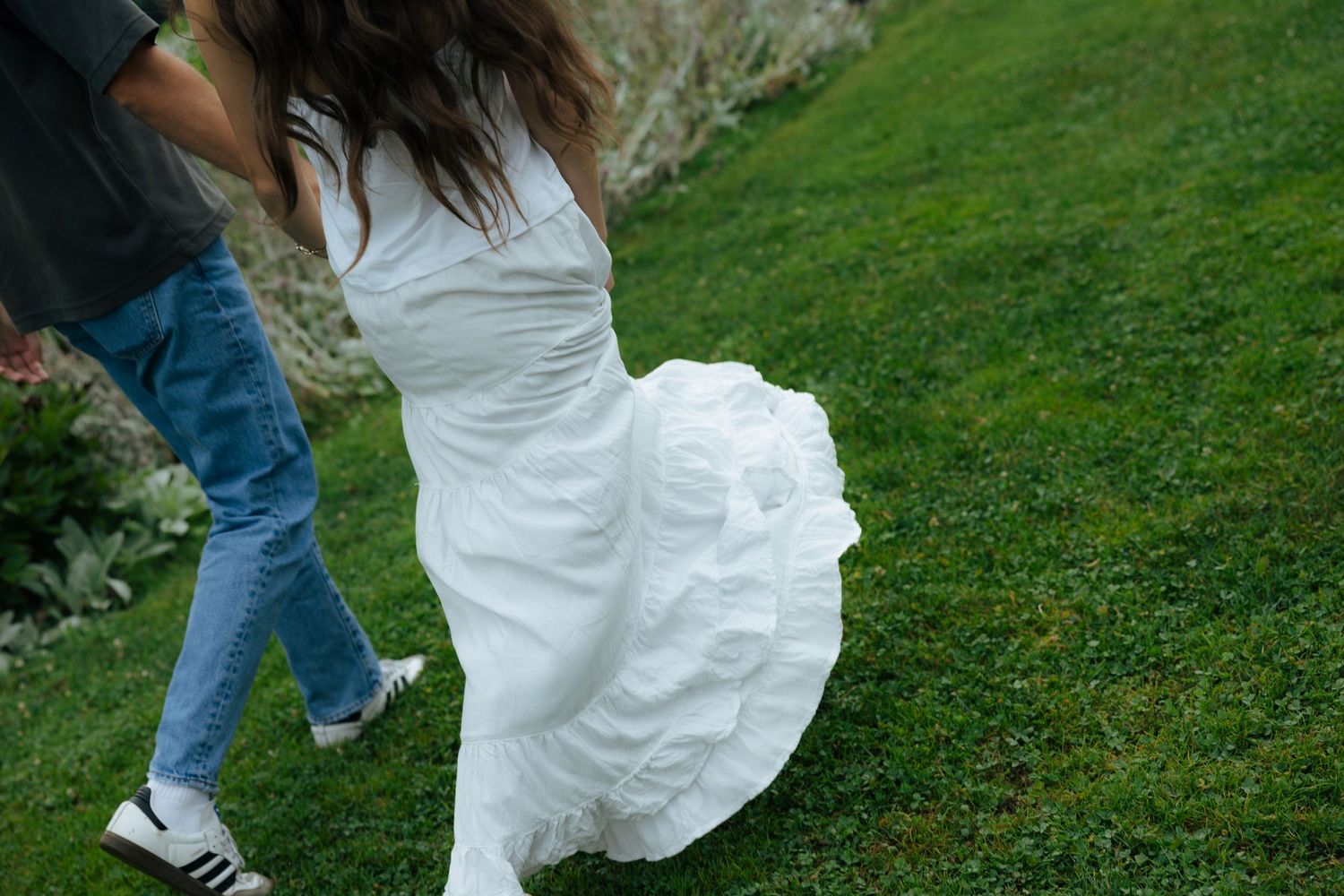 Bride in flowing white wedding dress walks on lush green grass with a guest in jeans and Adidas sneakers.