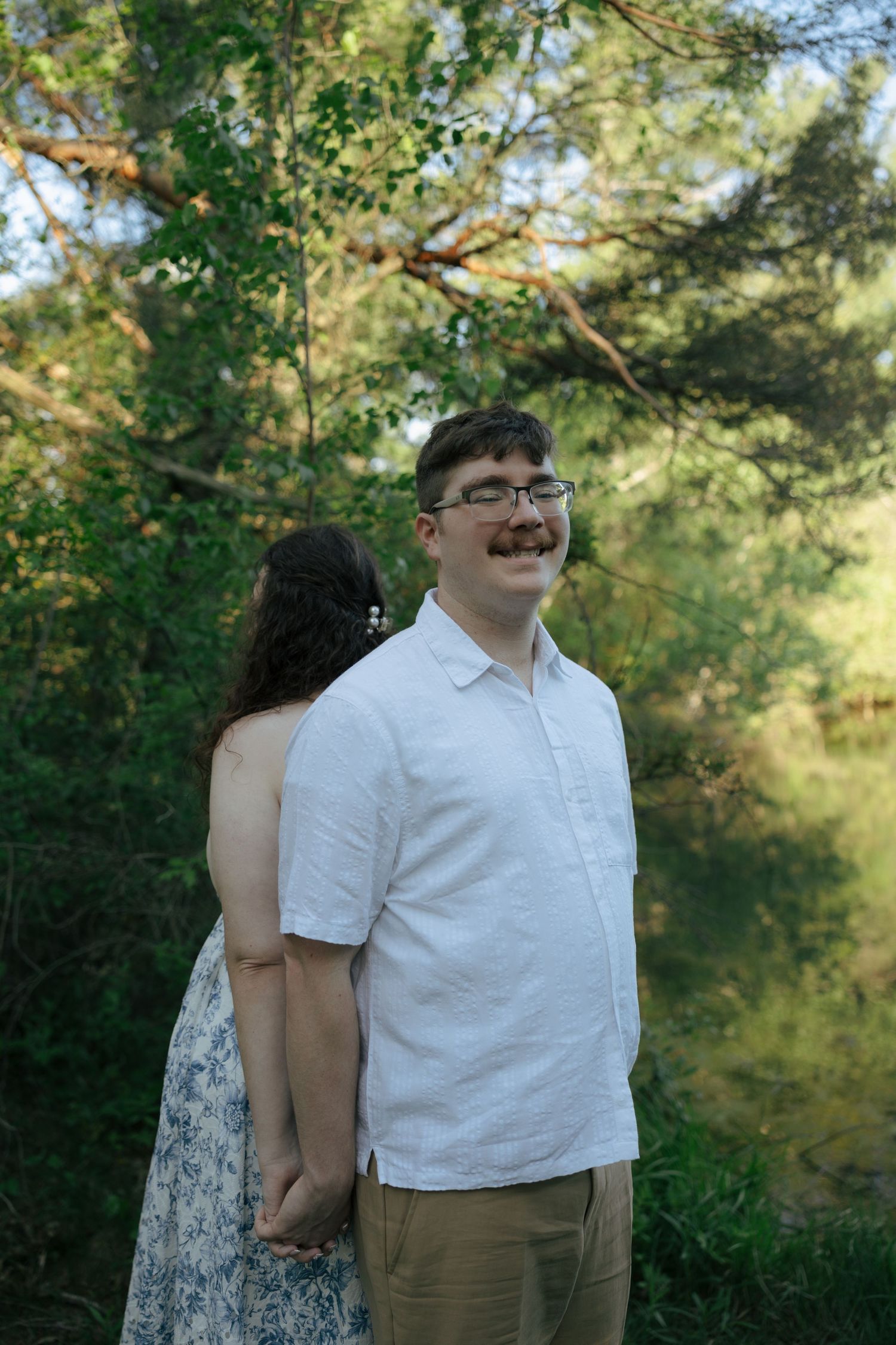 A smiling man with glasses and mustache in white shirt holds hands with a woman in floral dress outdoors.