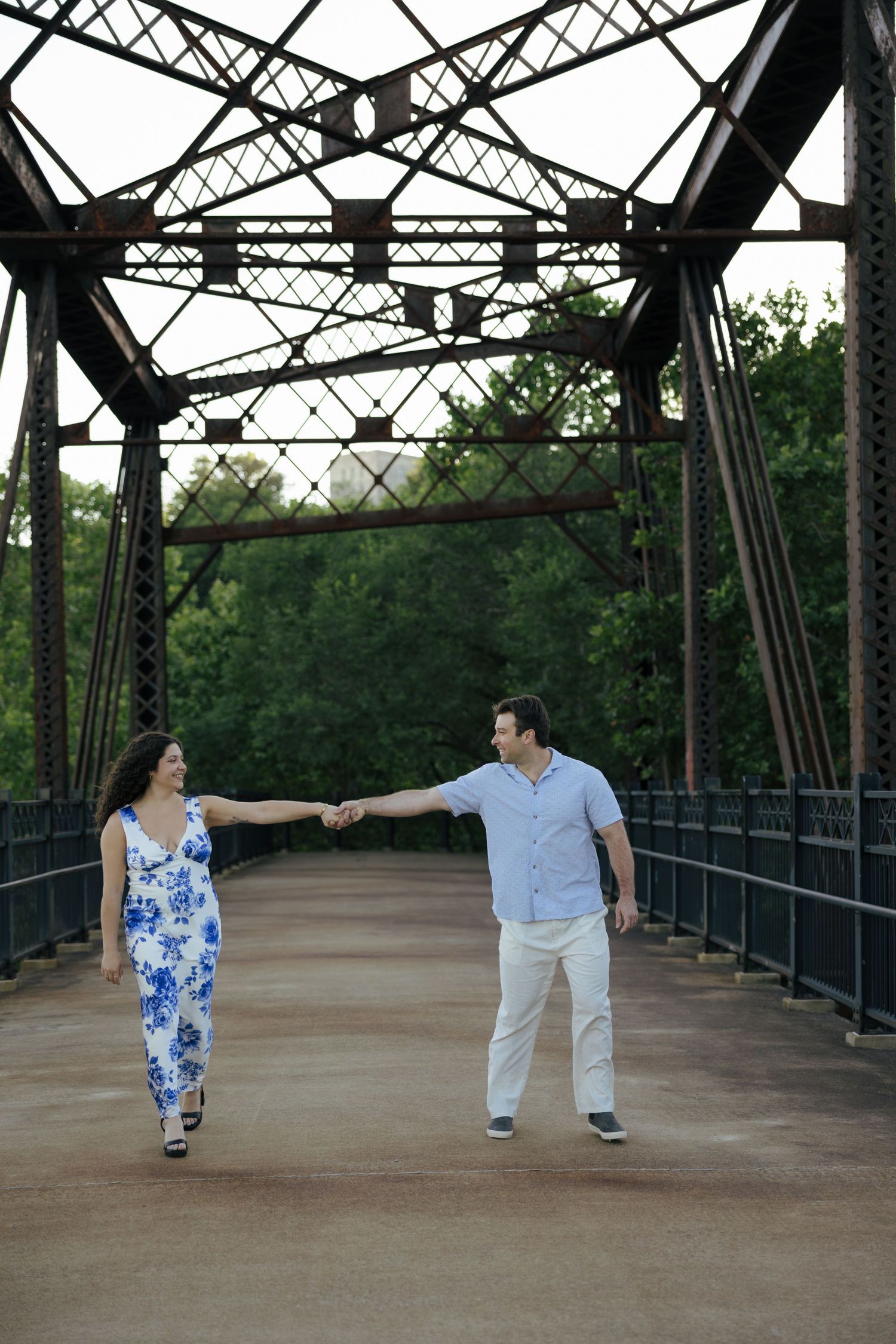 A couple holds hands and walks on a historic iron truss bridge surrounded by lush green trees.