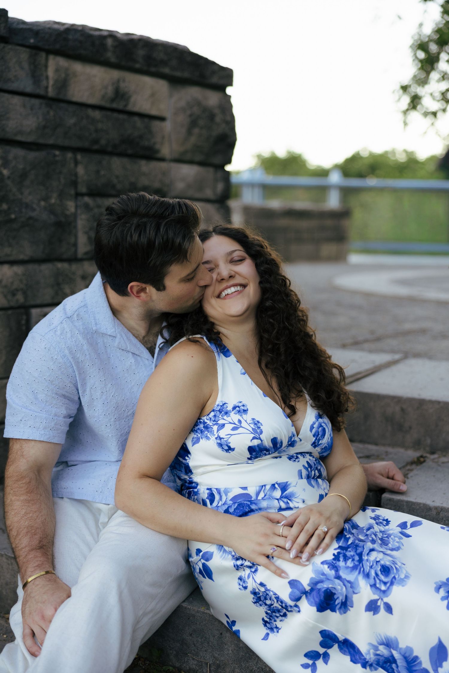 A smiling pregnant woman in a blue floral dress sits with her partner who kisses her cheek outdoors.