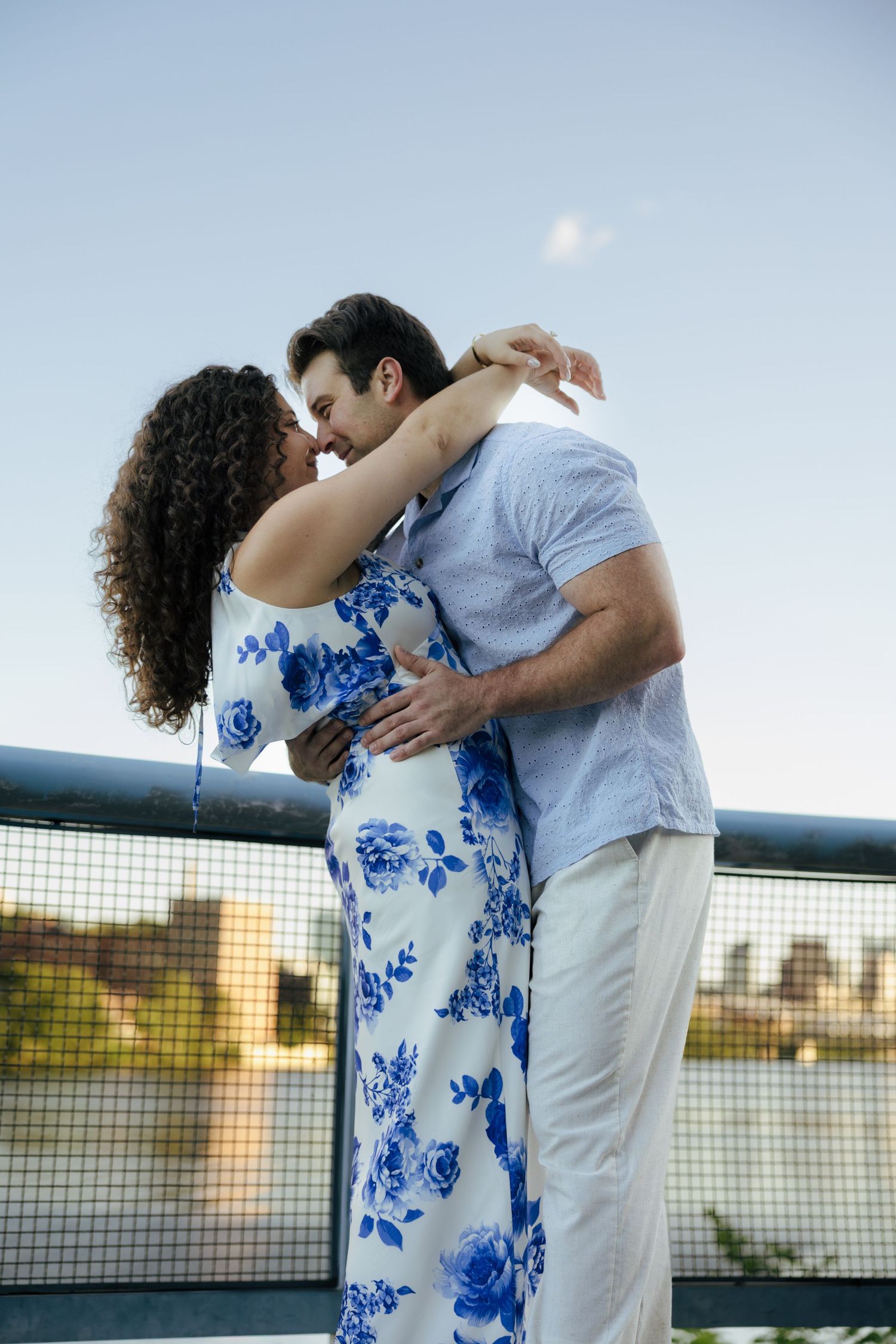 An engaged couple shares a romantic kiss on a bridge at sunset, woman in a blue floral dress.