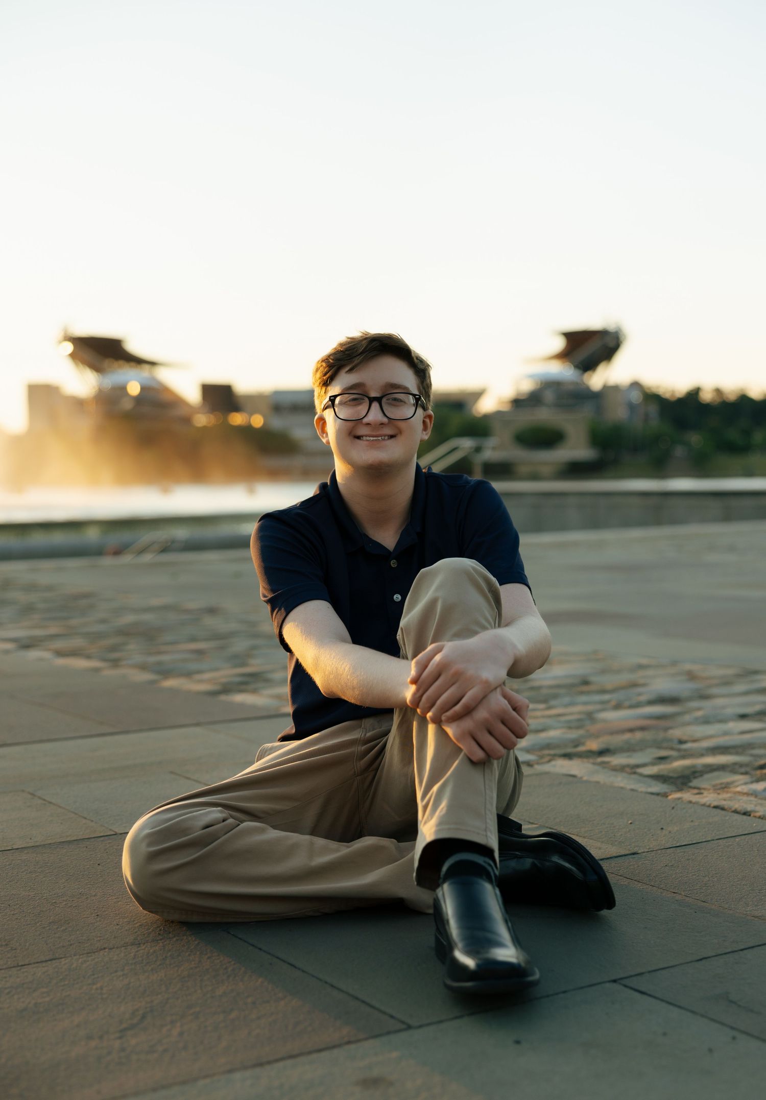 Smiling young person with glasses sitting cross-legged outdoors on pavement at sunset near a waterfront.