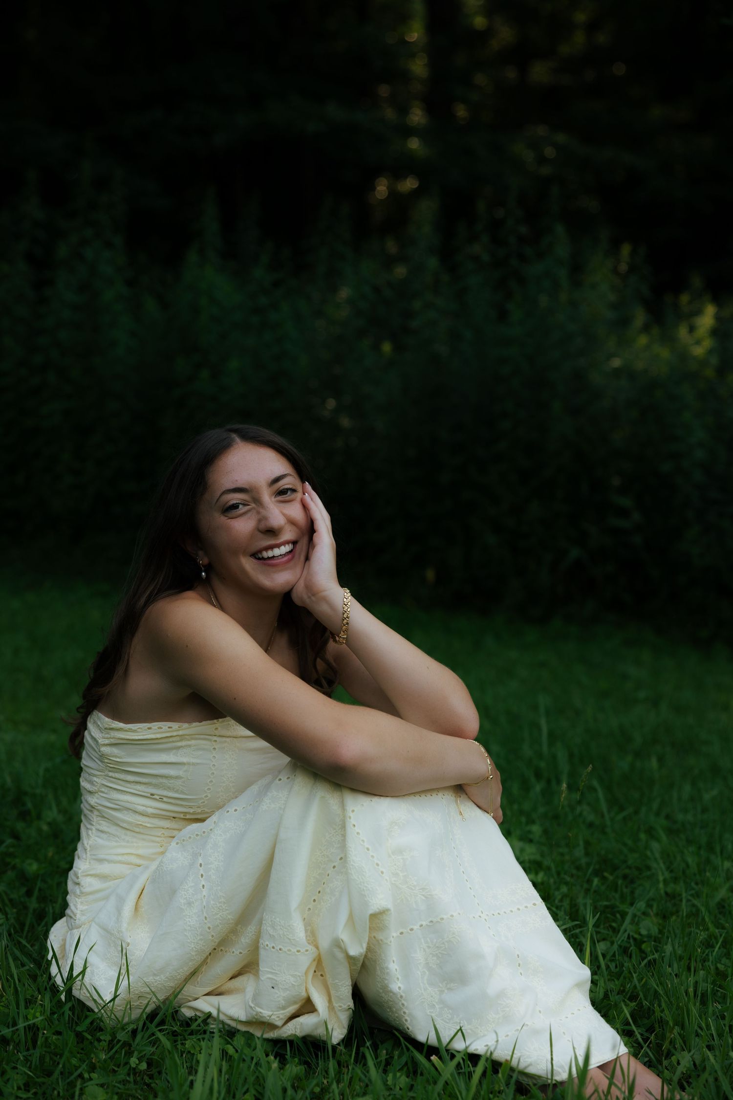Young woman in white dress sitting on grass, laughing joyfully with hand on cheek, green trees background.