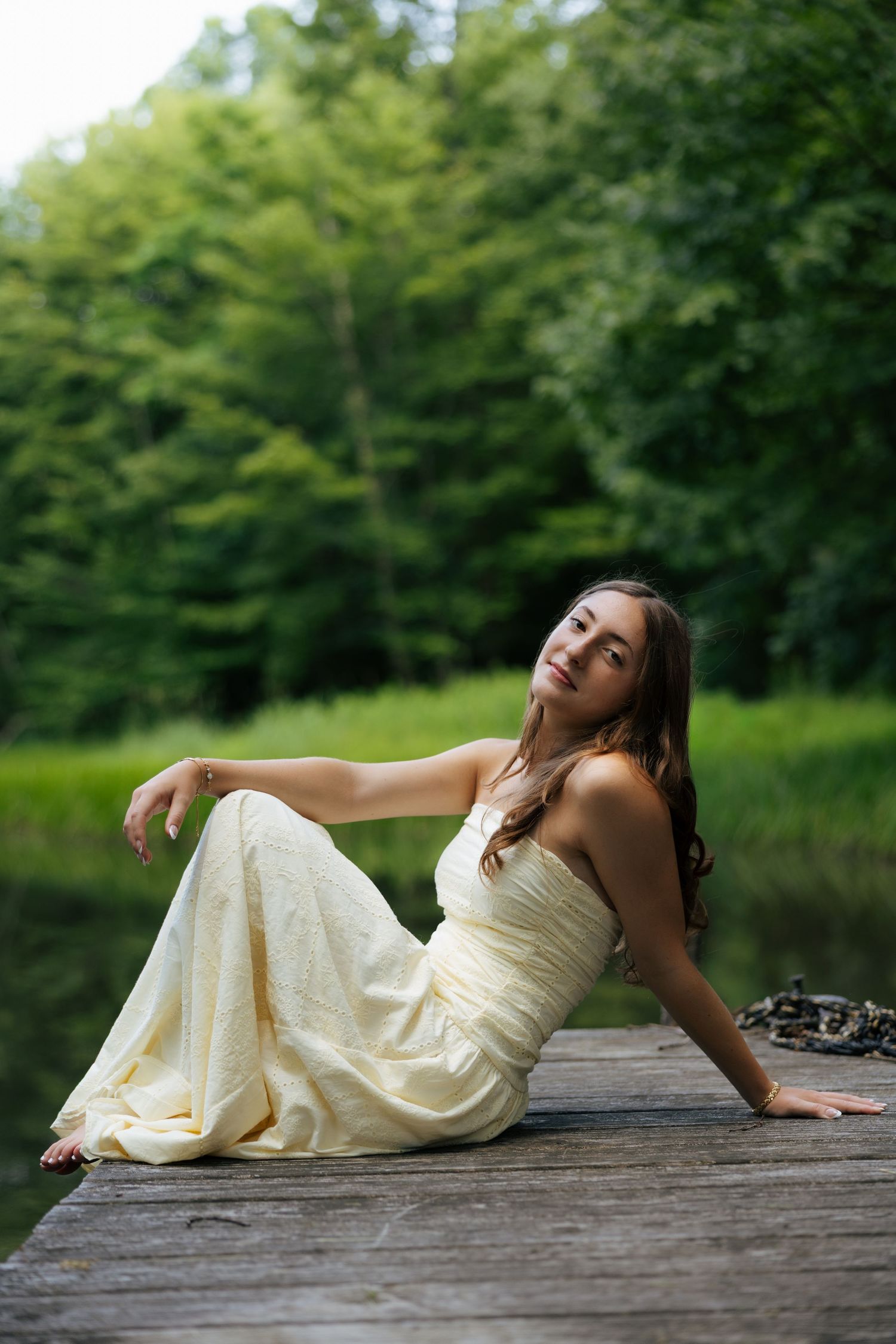 Young woman in flowing cream strapless gown sitting gracefully on wooden dock by forest lake.