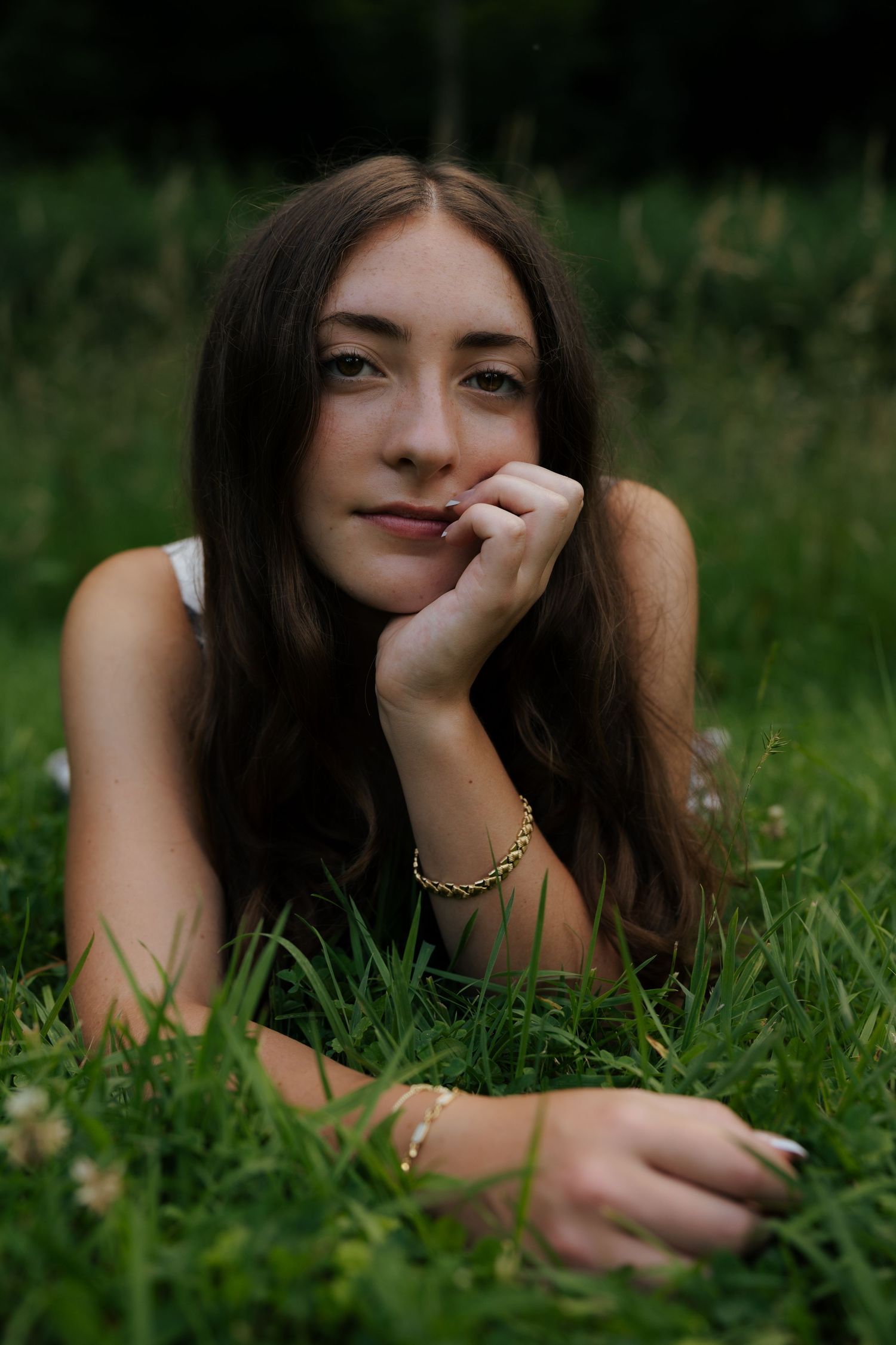 Young brunette woman lying in green grass, resting chin on hand, looking at camera outdoors.