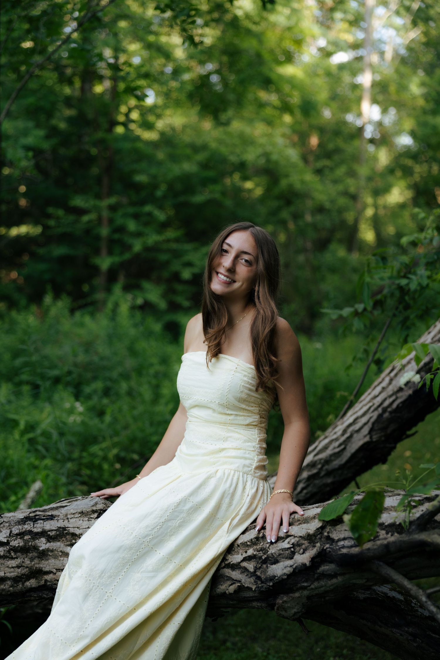 Young woman in yellow dress sitting on fallen log smiling in lush green forest.
