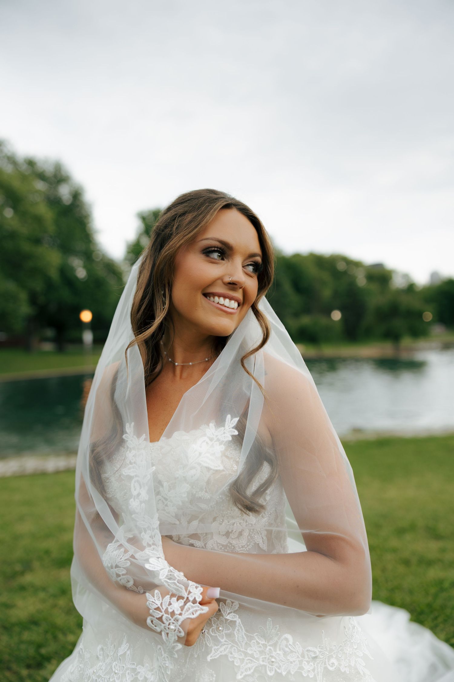 Smiling bride in white lace gown and veil poses outdoors near a pond on her wedding day.