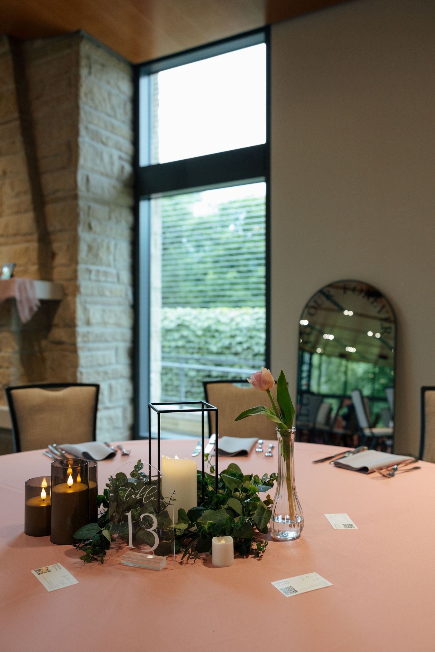 Elegant wedding table centerpiece with candles, greenery wreath, pink tulip, and table number 13 on a blush pink tablecloth.