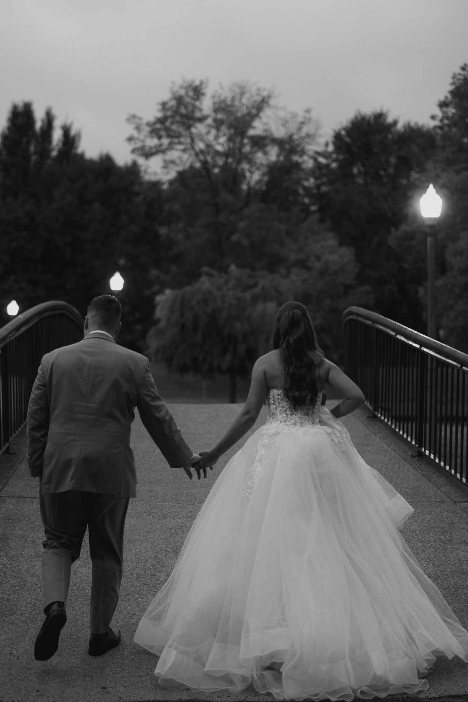 Bride and groom walk hand in hand across a bridge at dusk in a black and white wedding portrait.