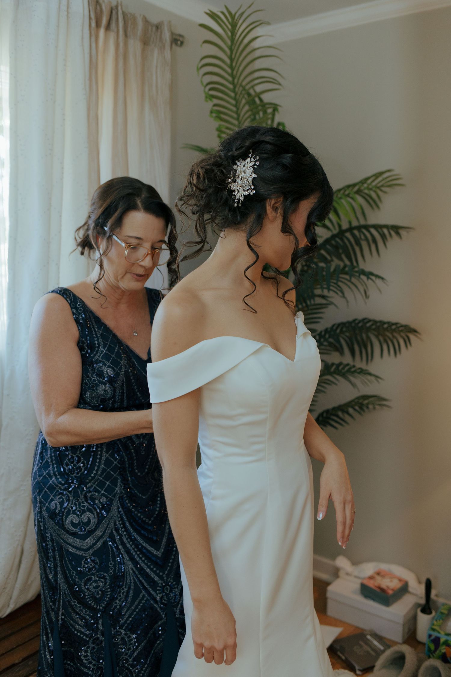Mother helps bride button up elegant off-shoulder white wedding gown in bright dressing room.