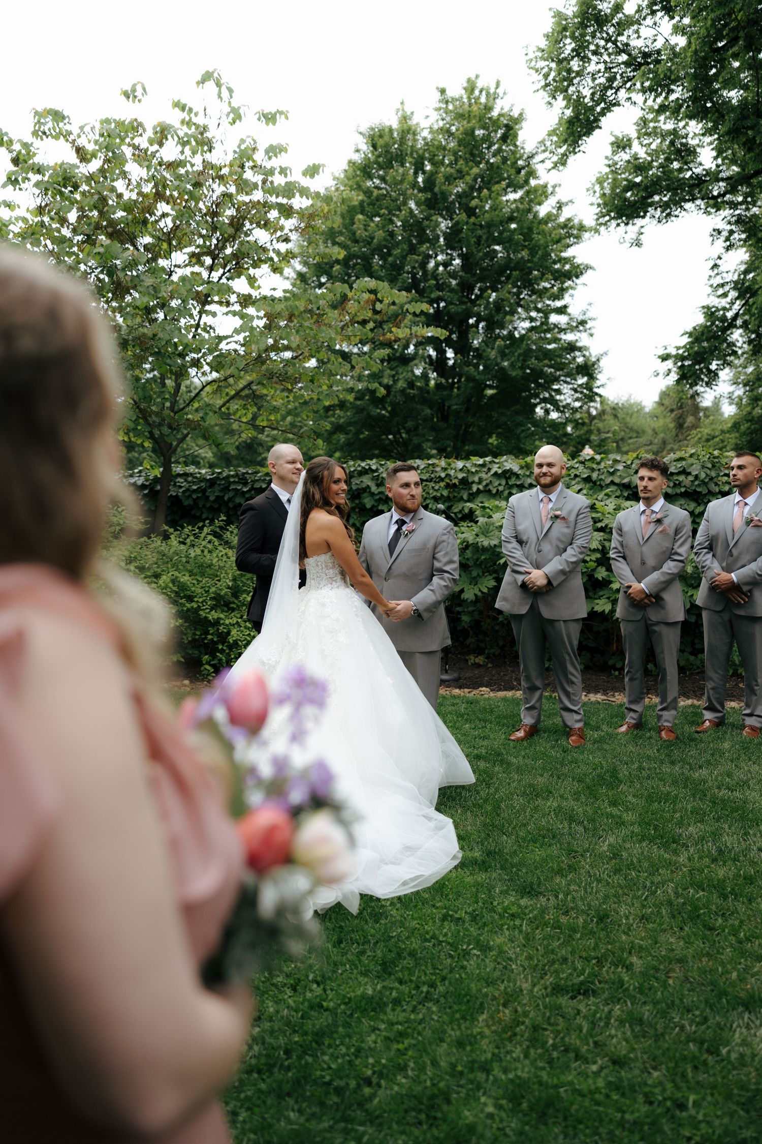 Bride in white gown stands with groom and groomsmen in gray suits during outdoor garden wedding ceremony.