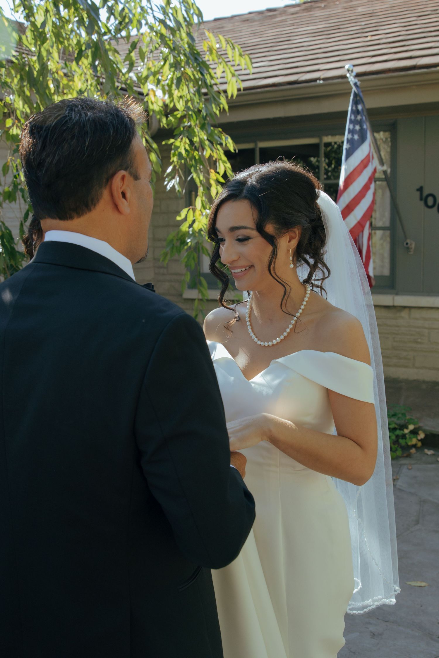 Bride in white off-shoulder gown and veil reads vows to groom outdoors, American flag visible behind them.