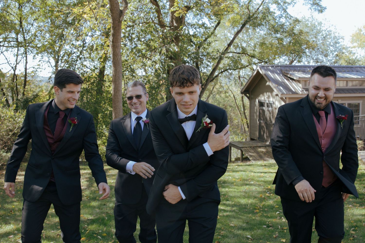 Four groomsmen in black suits with burgundy ties walk outdoors near rustic barn on a sunny wedding day.