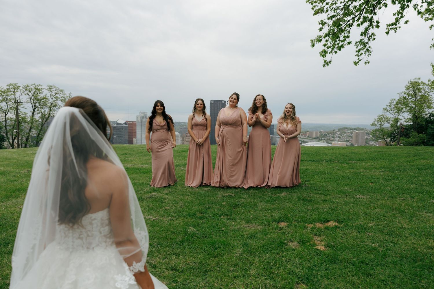 Bride in white gown and veil reveals herself to bridesmaids in dusty rose dresses on green lawn.