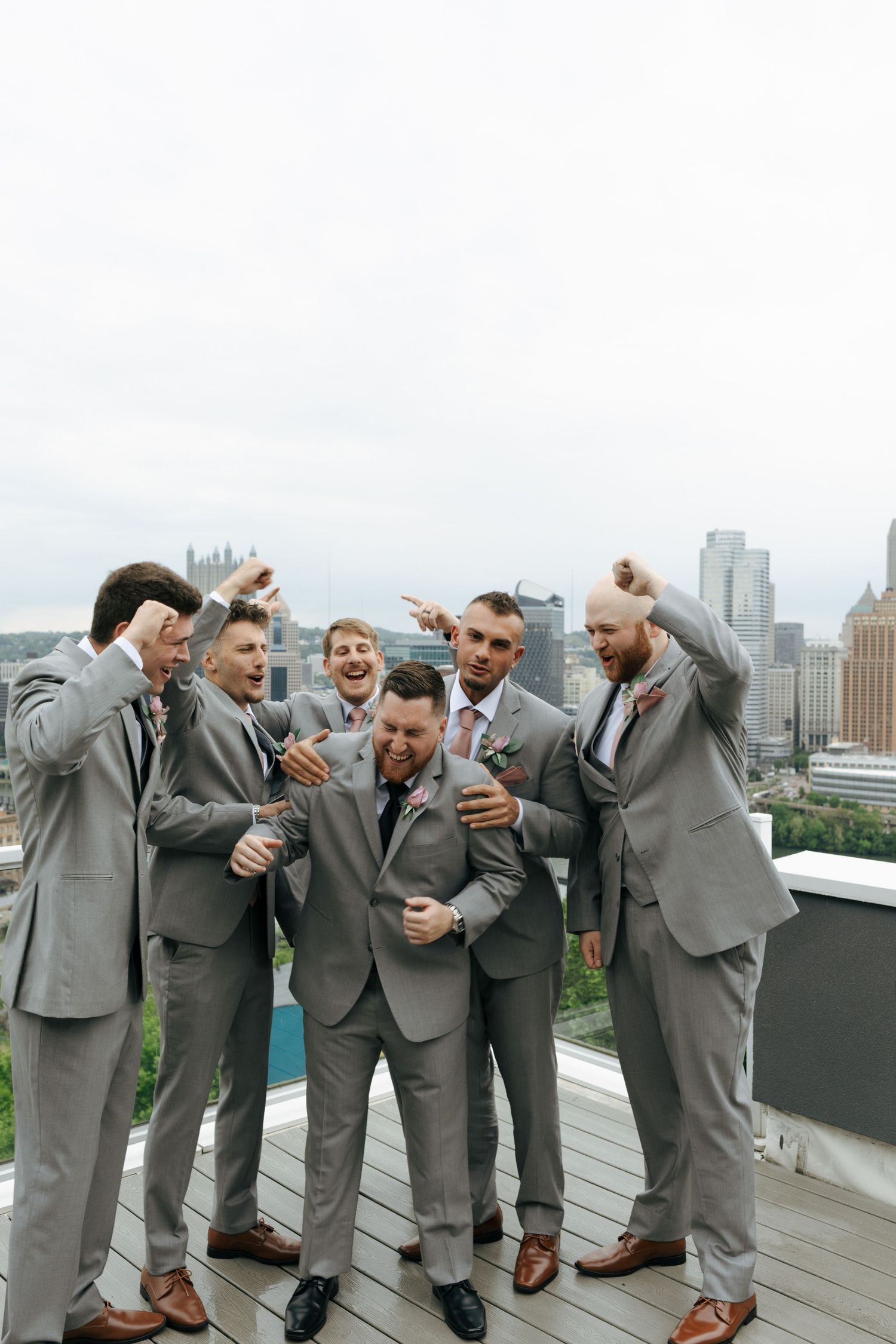 Groomsmen in grey suits celebrate on a rooftop with Pittsburgh skyline backdrop.
