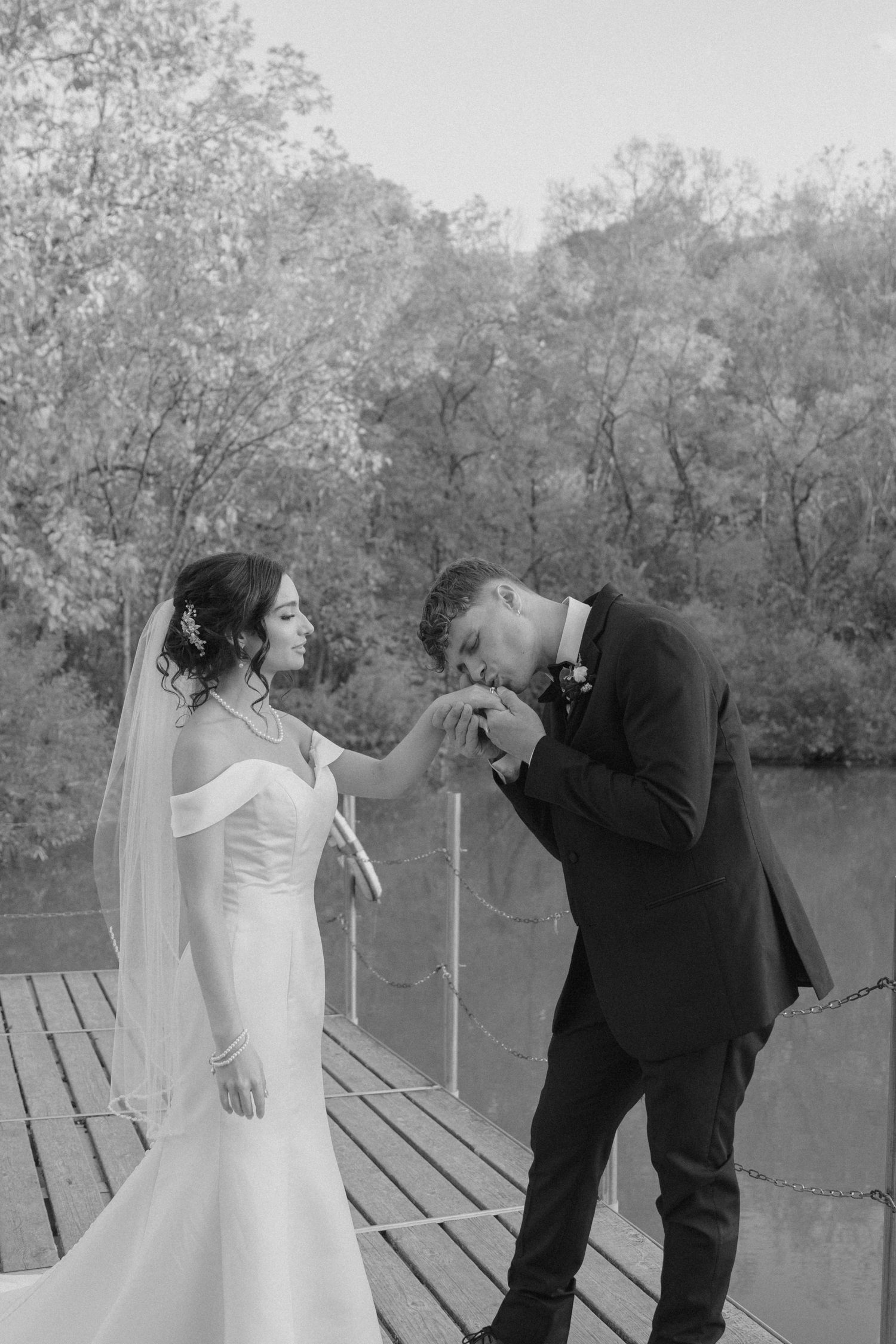 Black and white romantic photo of bride offering her hand to groom who bows to kiss it on a dock.