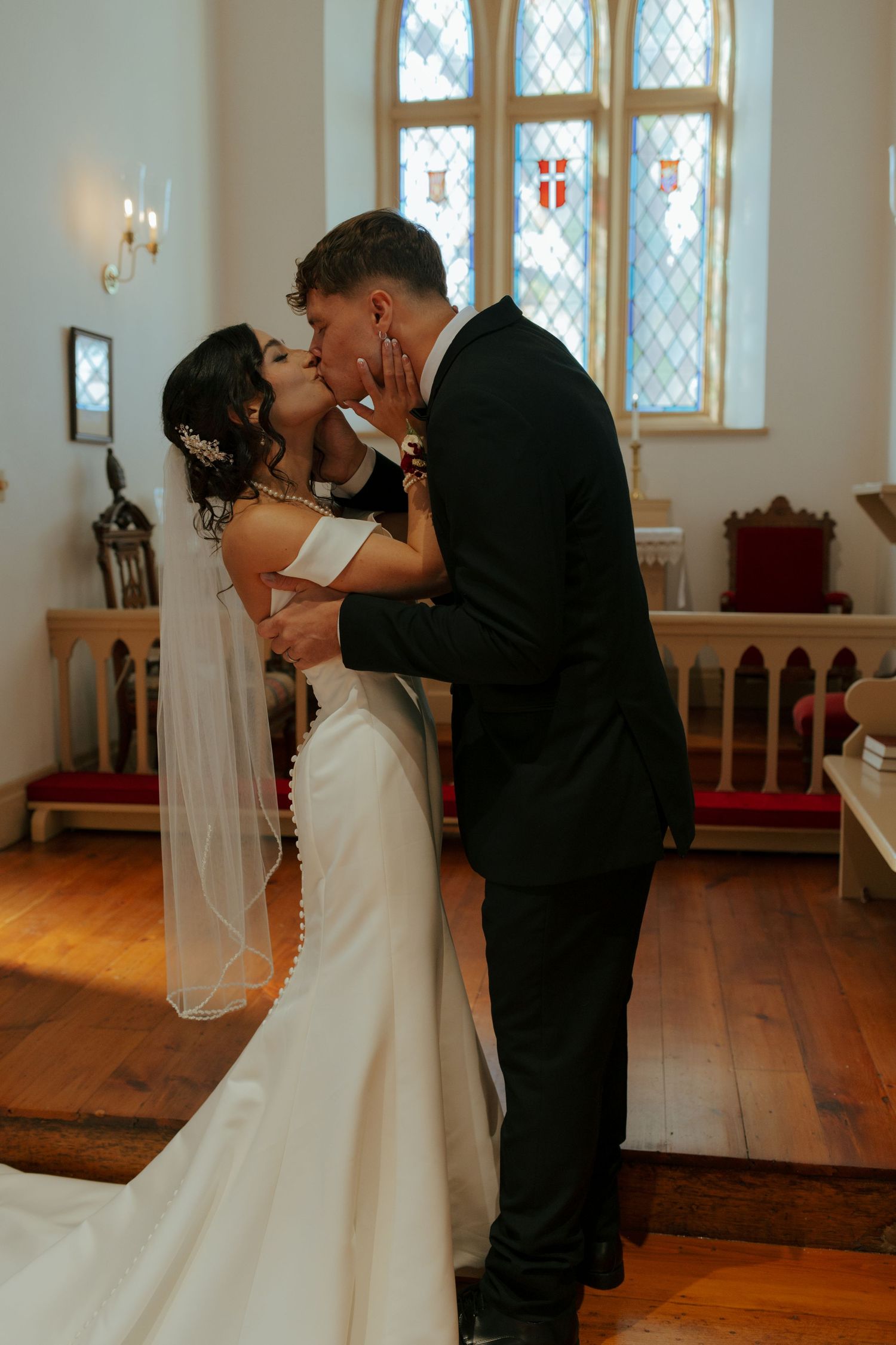 Bride and groom share their first kiss at the altar inside a beautiful historic church with stained glass windows.