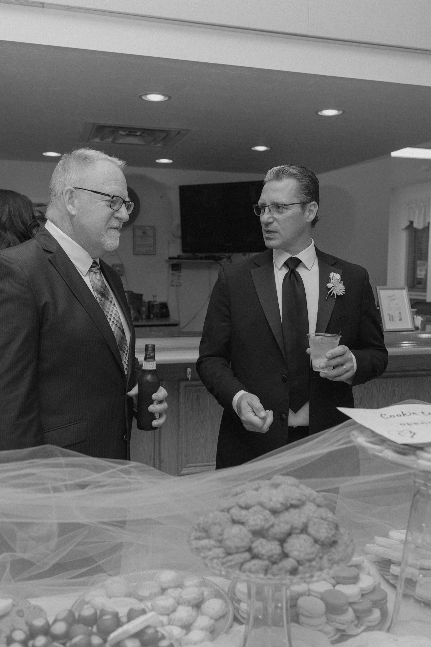 Black and white photo of two men in suits conversing near a dessert table at a wedding reception.