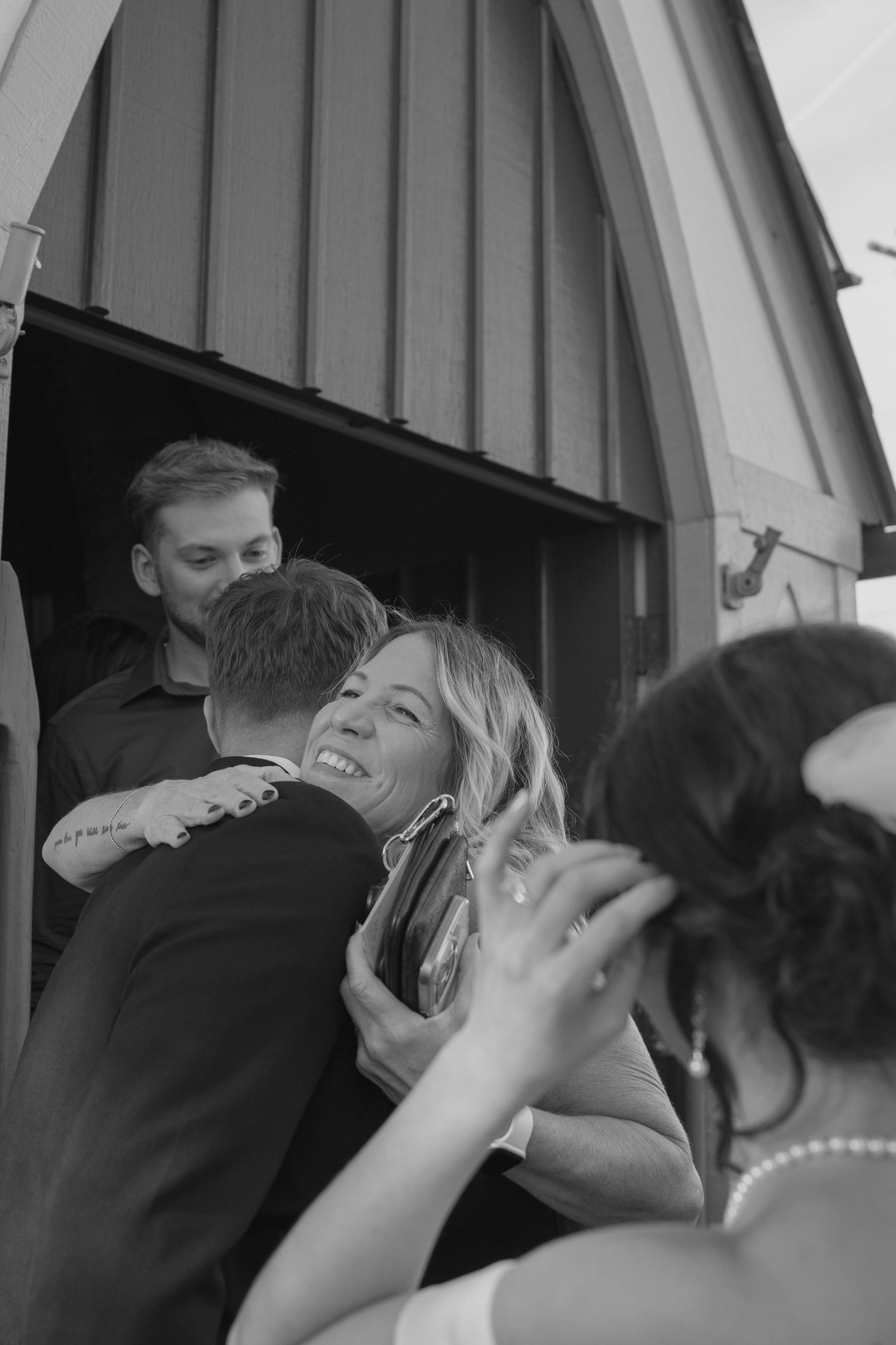 Black and white photo of a smiling woman hugging the groom at a rustic barn wedding celebration.